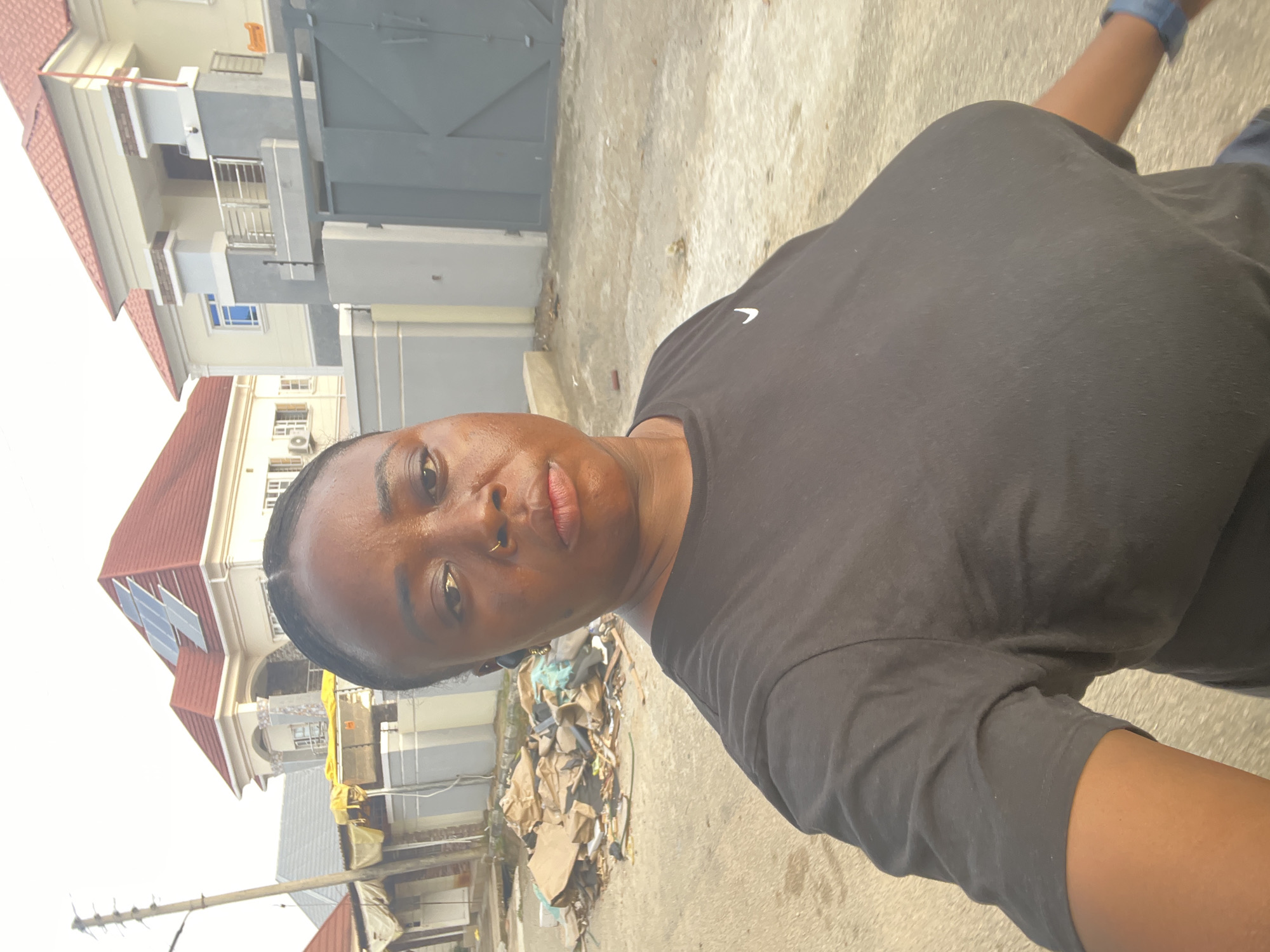 The image presents an individual taking a selfie in what appears to be a residential area. The person has dark hair, a gold nose ring, and is wearing a black t-shirt. The background features houses, one with solar panels on the roof, indicating possible modern construction. There is also visible trash and debris next to the path. The buildings and surrounding area suggest the location may be in Abuja, Nigeria, due to the architectural style and environmental elements common in that region. The area shows signs of mixed development, with both newer and more run-down elements present.