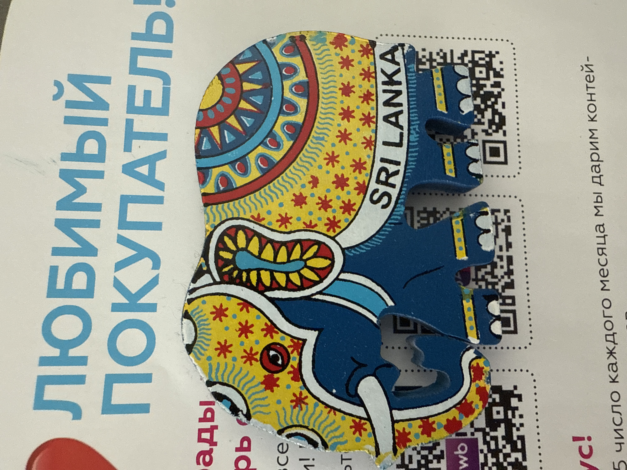 This media file is an image of a souvenir, likely a magnet or sticker, in the shape of an elephant with intricate patterns. The elephant is primarily blue with yellow, red, and black decorative elements. It is positioned on a white background that appears to be printed material.
The printed material has text in Russian, vertically oriented on the left side, which reads "ЛЮБИМЫЙ ПОКУПАТЕЛЬ!" (Beloved Buyer!). To the right of the elephant, there are two QR codes and more Russian text that appears to be promotional, stating "5 число каждого месяца мы дарим контей-" (On the 5th of every month, we give a container-). Another line of text reads "vc!" which could be part of a website or company name. On the elephant itself, the text "SRI LANKA" is clearly visible.
The overall impression is that this is a promotional item from Sri Lanka, likely distributed in Perm, Russia, to attract or thank customers. The bright colors and whimsical design of the elephant suggest a positive and cheerful theme. There are no people or animals present in the image itself, only the object and the printed material. The setting is a flat surface, likely a table or desk, given the lighting and the way the object is placed. There are no indicators of time of day or weather.