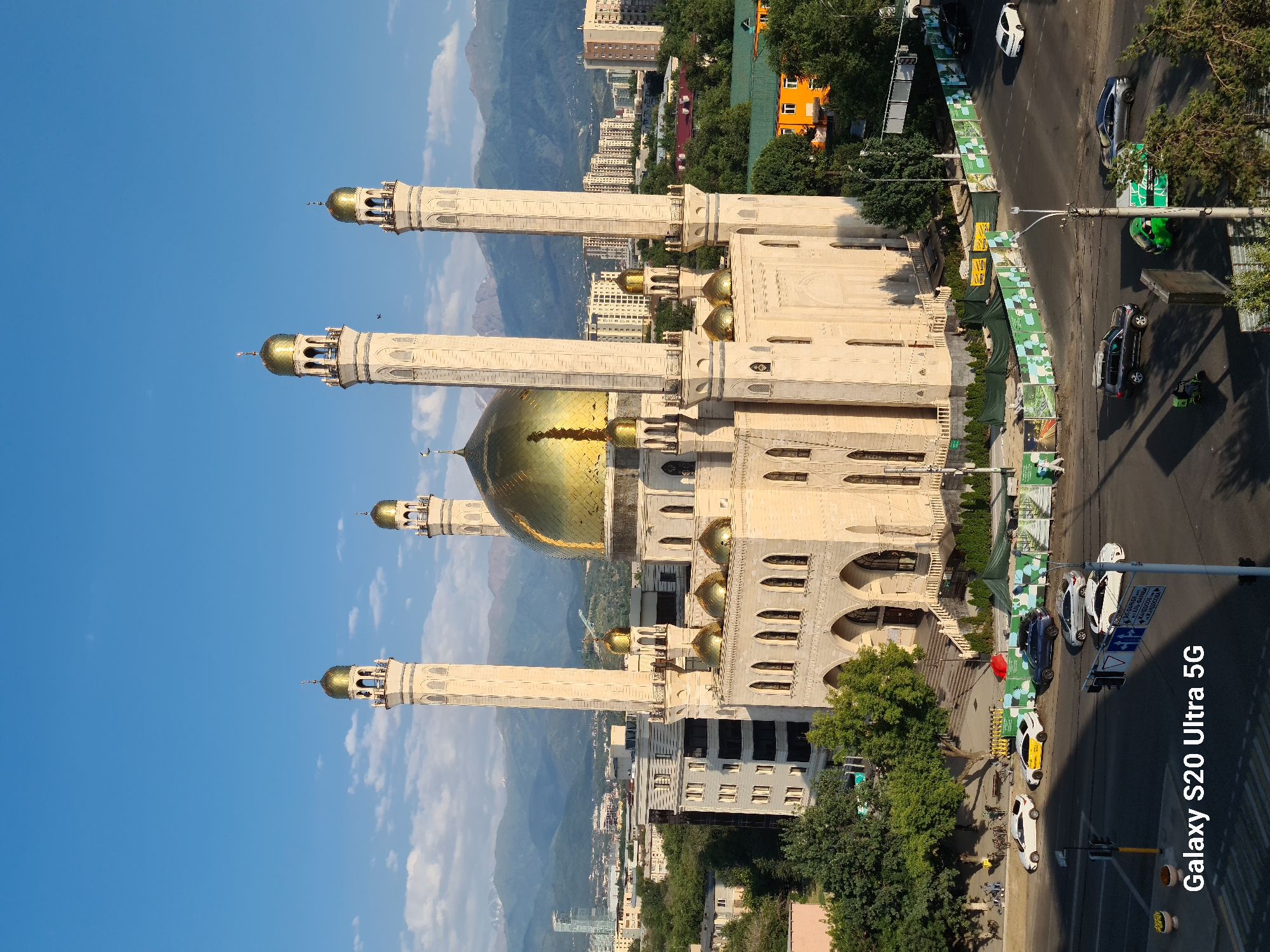 The image captures a wide-angle view of the Central Mosque in Almaty, Kazakhstan, under a clear blue sky with scattered clouds. The mosque, with its prominent golden dome and four tall minarets, is the central subject, bathed in sunlight. The architectural style is characteristic of Islamic design, featuring intricate stonework and multiple smaller golden domes.
Surrounding the mosque is a mix of urban and natural elements. In the foreground, a busy intersection with a multi-lane road is visible, with several cars, including a white sedan, a dark SUV, and a green vehicle, navigating the traffic. Traffic lights and street signs are present. Adjacent to the road, pedestrian walkways and greenery can be seen, with some trees providing shade. There are also construction barriers adorned with informational graphics along the perimeter of the mosque grounds.
In the middle ground, several multi-story buildings, ranging in architectural style, are interspersed with trees. Further in the background, a range of mountains, possibly part of the Tien Shan range, forms a majestic backdrop against the skyline, their peaks dusted with snow. The overall scene depicts a blend of religious and modern urban life within a picturesque natural setting. The image is captured with a Samsung Galaxy S20 Ultra 5G phone, as indicated by the watermark in the bottom left corner.