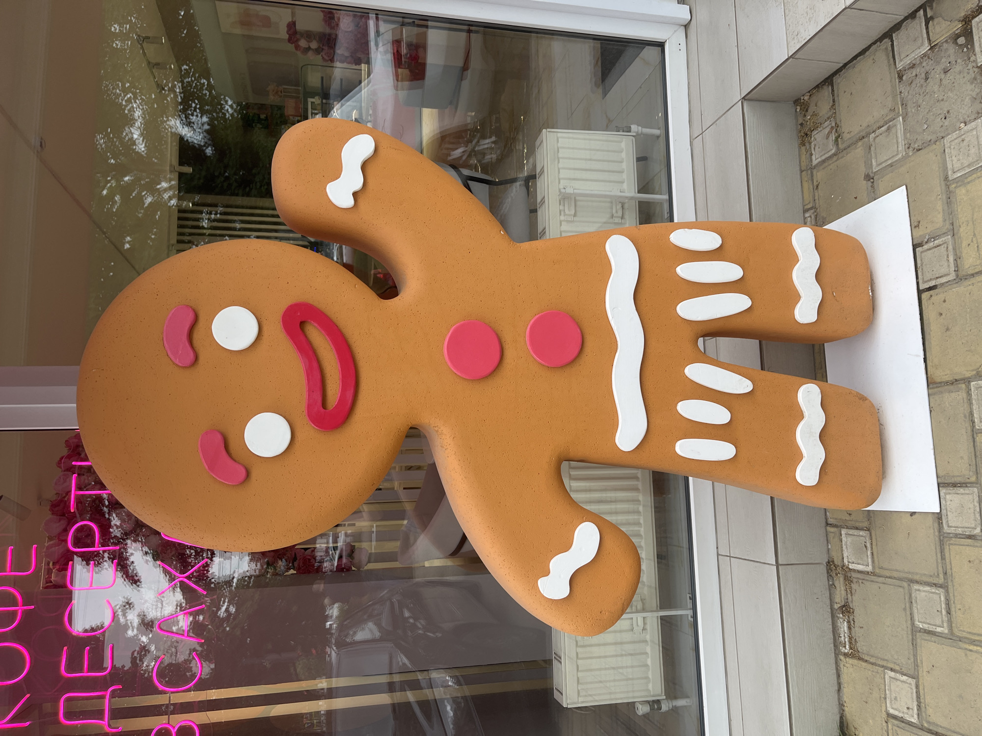 A large, stylized gingerbread man decoration stands outside a window of a business in what appears to be an urban area. The gingerbread man is light brown with white and red decorative elements resembling icing. The decorations on the gingerbread man include circles for buttons, and a curved line for a mouth. The gingerbread man is standing on a white board on the sidewalk. The window behind the gingerbread man reflects the surroundings, with hints of pink neon signage. The sidewalk is paved with square stones. The location is Krasnodar, Russia.