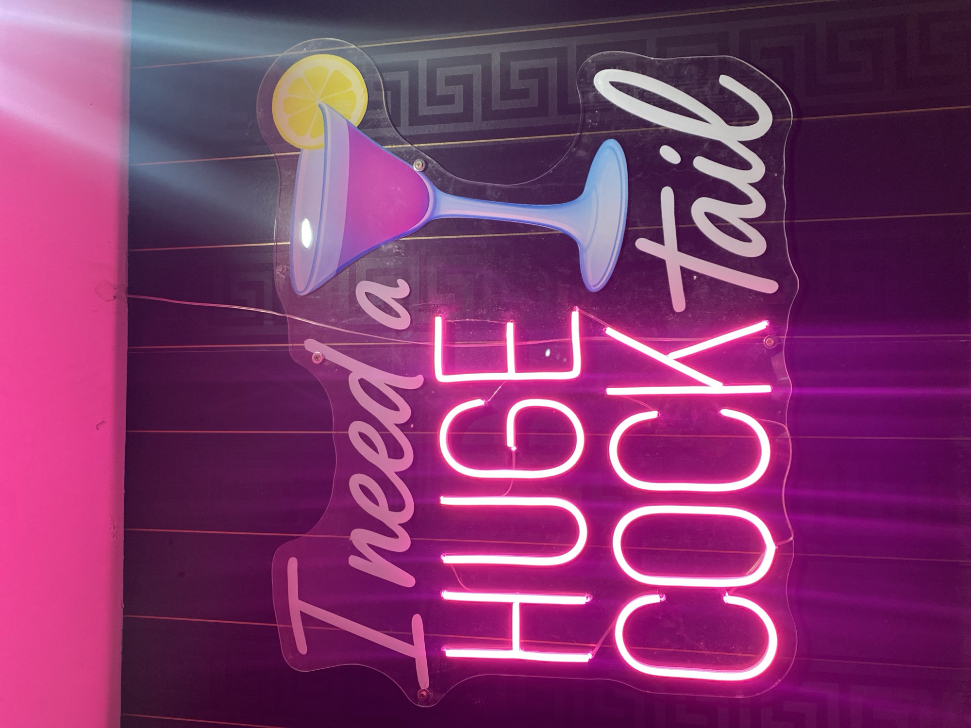 A vertically oriented, close-up shot shows a lit neon sign. The sign is transparent and shaped somewhat like a cocktail glass. A cartoon cocktail glass is depicted at the top of the sign with a martini-style glass filled with pink liquid and garnished with a lemon slice. The neon light highlights the phrase "I need a HUGE COCKtail" in pink. The background is a dark, horizontally striped surface, possibly a wall, with a Greek key pattern subtly visible. A bright pink vertical surface is visible on the left edge of the frame. The location in the city of London, United Kingdom is not evident from the image.