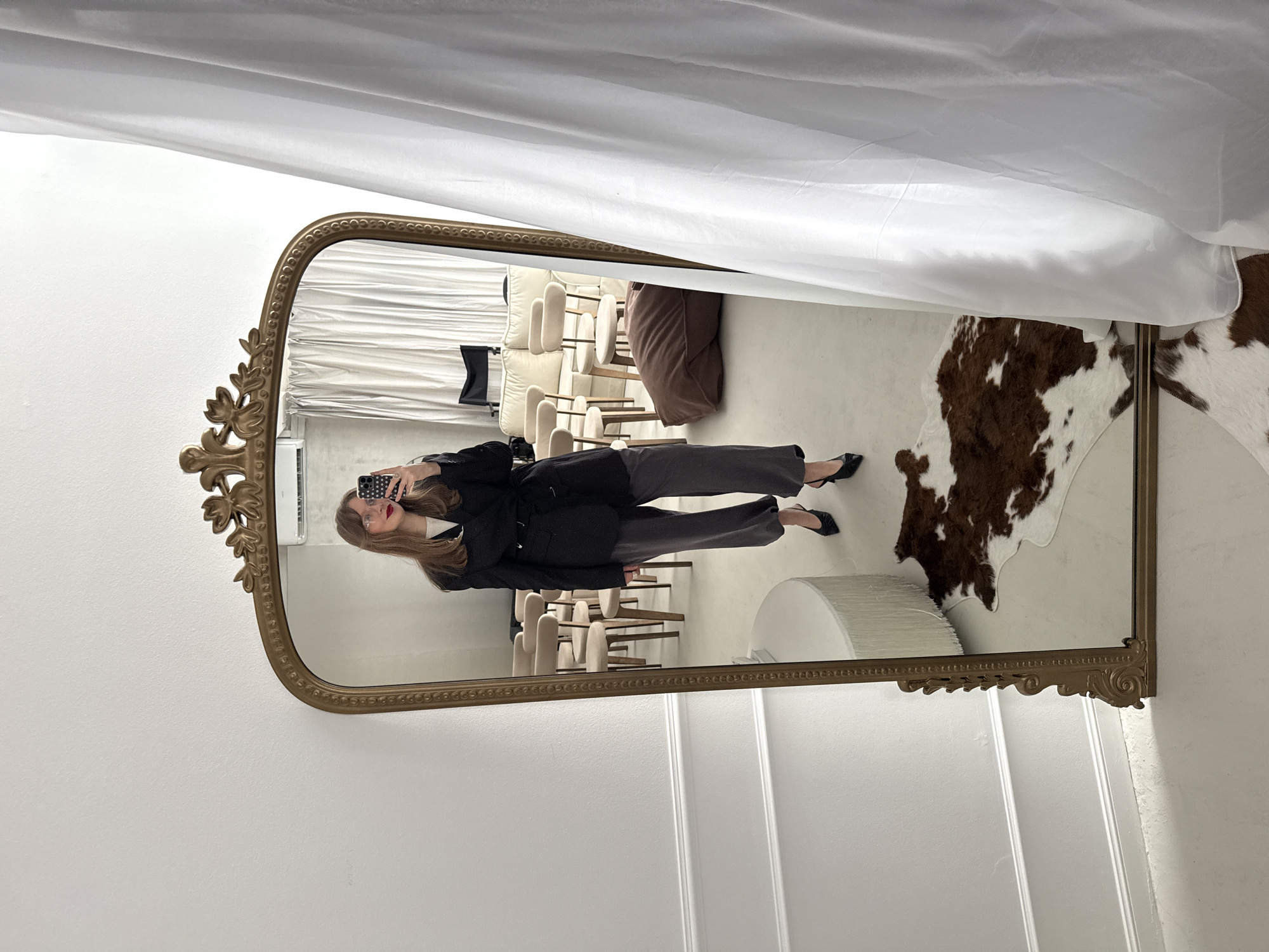 A mirror reflects a woman taking a photo. She is wearing a black blazer, gray pants, and black heels. The setting includes decorative white drapes at the top, a faux cowhide rug, a modern white table lamp, and several chairs arranged in the background. The surrounding environment appears to be an indoor space with white walls and decorative molding on the lower section. There is no information that suggests the image was taken in Izhevsk, Russia.
