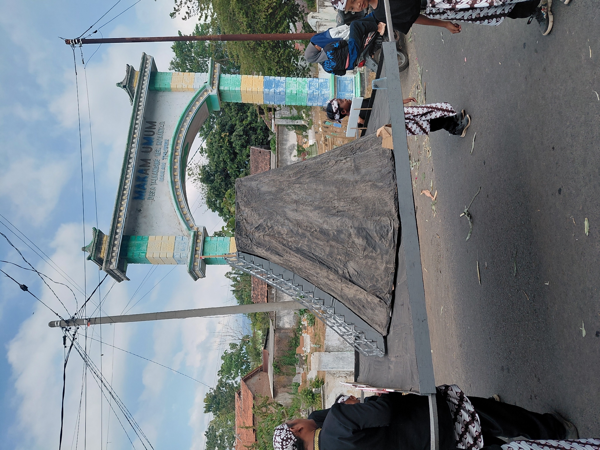 The image depicts a street scene in Bimo, Indonesia, in front of an archway labeled "MAKAM UMUM DESA TALIKDANG KEC. KOTAANYAR MAKAM THUWET". The archway is decorated with green, yellow, and blue colors. In the foreground, a large, dark-colored conical structure covered with tarp is set up on a platform. A staircase with a silver railing is attached to the side of the structure. Several people are visible around the platform. On the left, a man in traditional Indonesian attire, including a black jacket and a patterned sarong, is facing away from the camera. To his right, a young person in a similar sarong is partially visible. Further to the right, a few children in traditional attire are present, and a woman on a motorcycle is also visible in the background, to the right of the conical structure. The ground is paved, and there are some scattered leaves and debris. Beyond the platform and archway, there are houses with red tile roofs and some trees, suggesting a rural or semi-urban setting. The sky is partly cloudy. The overall scene appears to be part of a cultural event or ceremony.