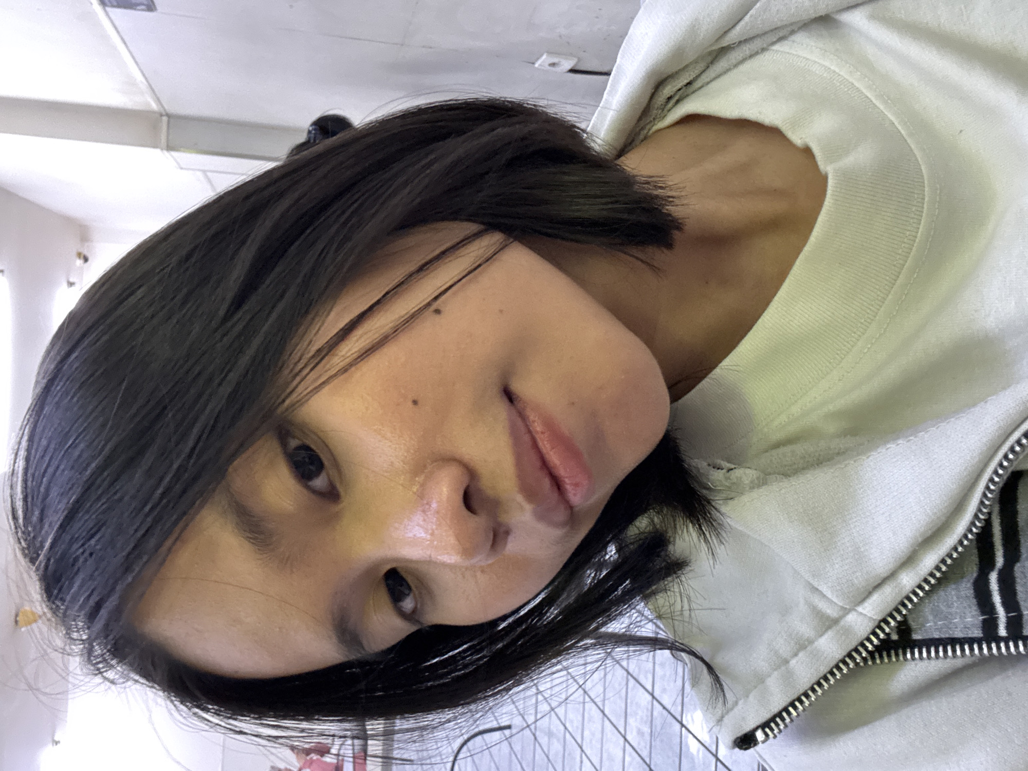 A young woman with shoulder-length black hair takes a selfie. She is wearing a white t-shirt and a light-colored zip-up hoodie. Her head is tilted slightly to her left, and she is looking directly at the camera. The background appears to be an indoor setting with tiled flooring and a light-colored wall. A light switch is visible on the wall. The lighting suggests an indoor environment. Based on the context of East Jakarta, Indonesia, the location could be a public space like a mall, cafeteria, or an indoor common area.