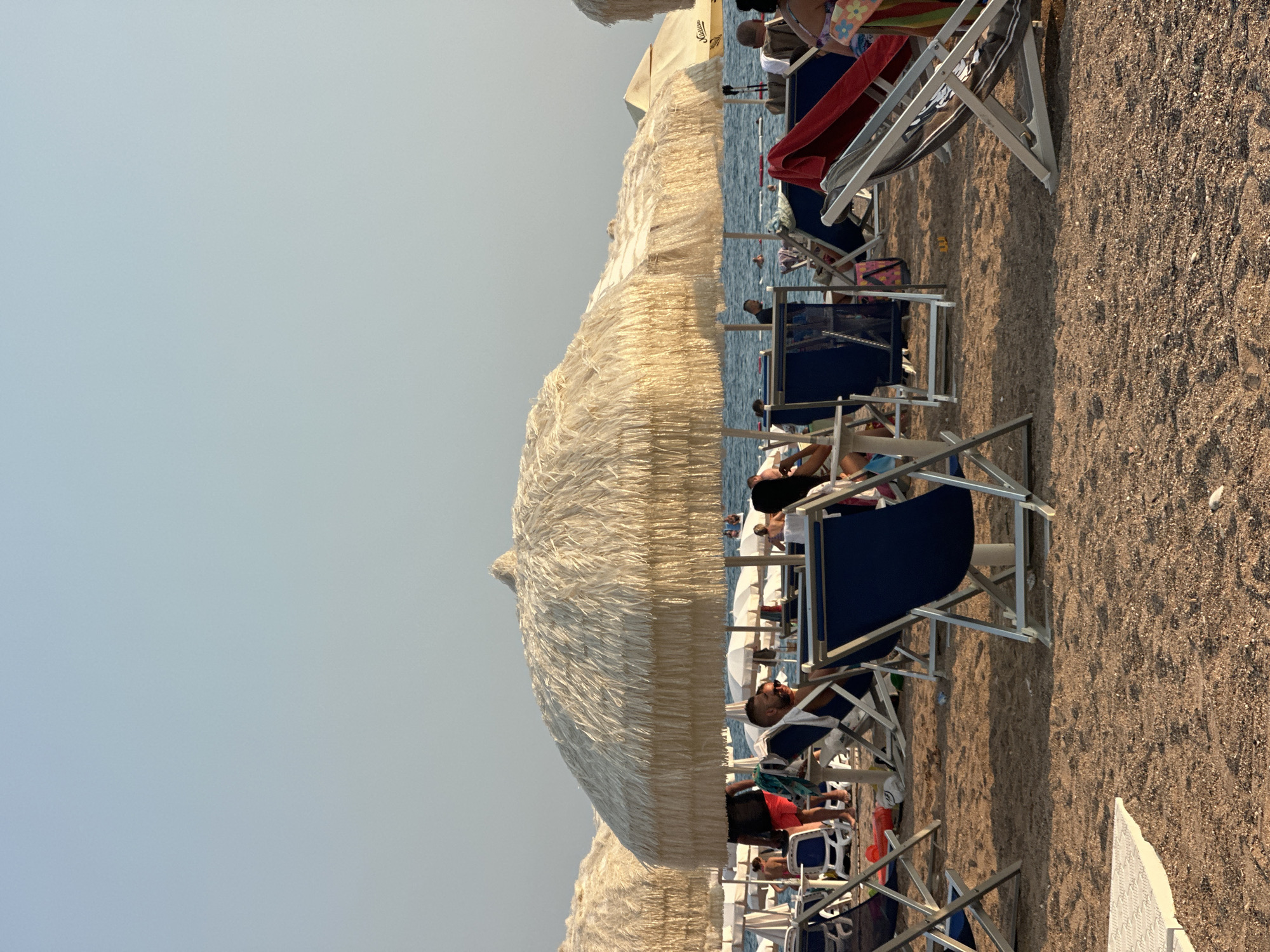 The image depicts a crowded beach scene in Boreti, Montenegro, with a focus on several straw-thatched umbrellas providing shade over rows of lounge chairs on the sandy shore. In the foreground, several blue and white lounge chairs are visible, some occupied by individuals. A man wearing red shorts and a black t-shirt is walking away from the camera, towards the water. Further back, more lounge chairs and umbrellas are spread across the beach, with numerous people visible in the distance, some swimming or wading in the calm, blue sea under a clear, pale sky. The overall atmosphere suggests a typical summer day at a popular seaside resort.