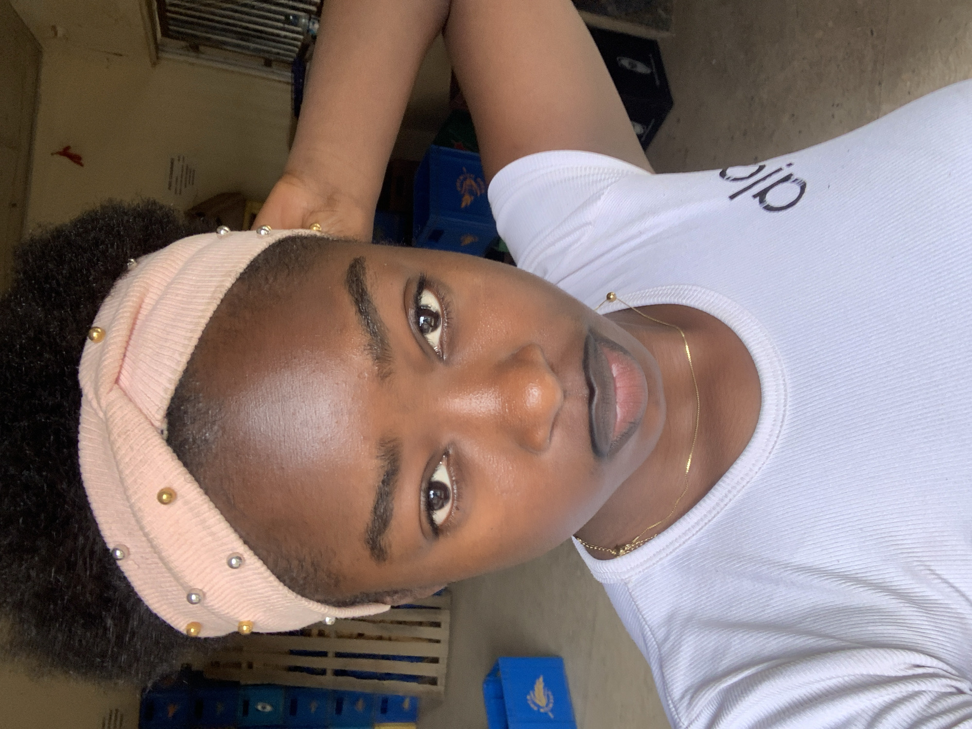 A close-up, medium shot of a young, Black woman with dark hair, wearing a light pink headband with gold and silver beads, and a white shirt with the word "alo" on it, lies on her back in what appears to be a casual indoor setting. She has a thin gold necklace around her neck. Her gaze is directed at the camera. The location may be New Stadium, Nigeria, based on the request, but there is no visible evidence in the image to confirm this. Visible in the background are blue plastic crates, a window, and some vague interior features, but the background is generally out of focus and does not offer specific location information. No actions or interactions are depicted beyond the woman's pose.