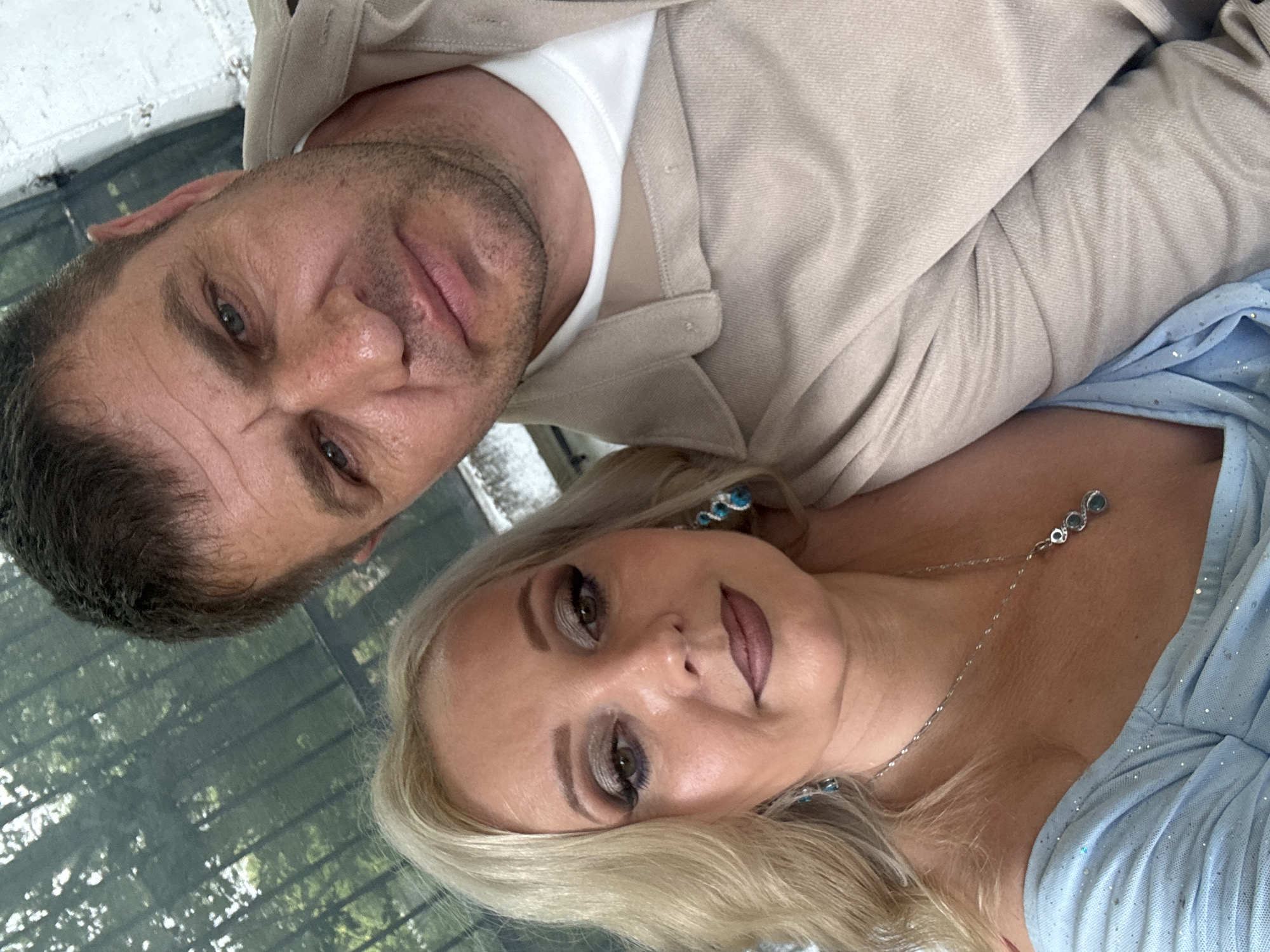 A close-up, slightly angled selfie features a man and a woman positioned in the foreground. The man, with short dark hair, is on the right, his face turned towards the camera, displaying a neutral expression. He is wearing a light beige collared shirt over a white t-shirt. The woman, on the left, has blonde wavy hair and is looking directly at the camera with heavy eye makeup and a matte lipstick. She is wearing a light blue off-the-shoulder dress adorned with sparkles and a silver necklace with a pendant. Blue earrings are visible on both her ears. The background appears to be an outdoor setting with a dark green mesh-like covering overhead, through which trees are visible, suggesting a shaded area, possibly a park or a pergola. The overall lighting is natural. This image was taken in Komsomolsk-on-Amur, Russia.