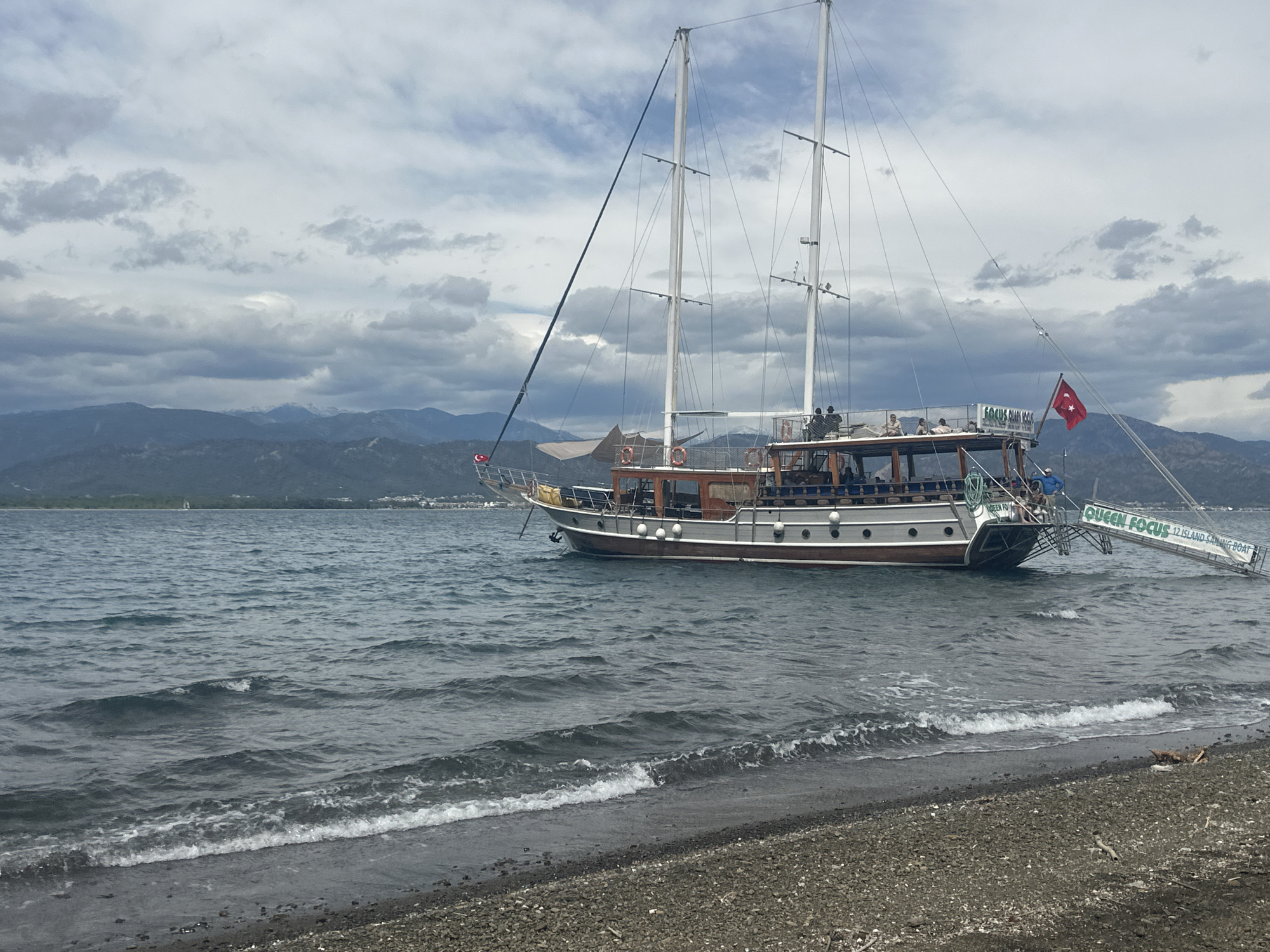The image depicts a large, two-masted sailing vessel, named "Queen Focus 12 Island Sailing Boat", close to a shoreline in Fethiye, Turkey. The boat is anchored near a beach composed of dark pebbles. A gangway extends from the stern of the boat, reaching towards the shore. People can be seen on the boat's deck. The Turkish flag flies from the stern of the vessel. The water is choppy with small waves lapping against the shore. In the background, mountains are visible under a cloudy sky.