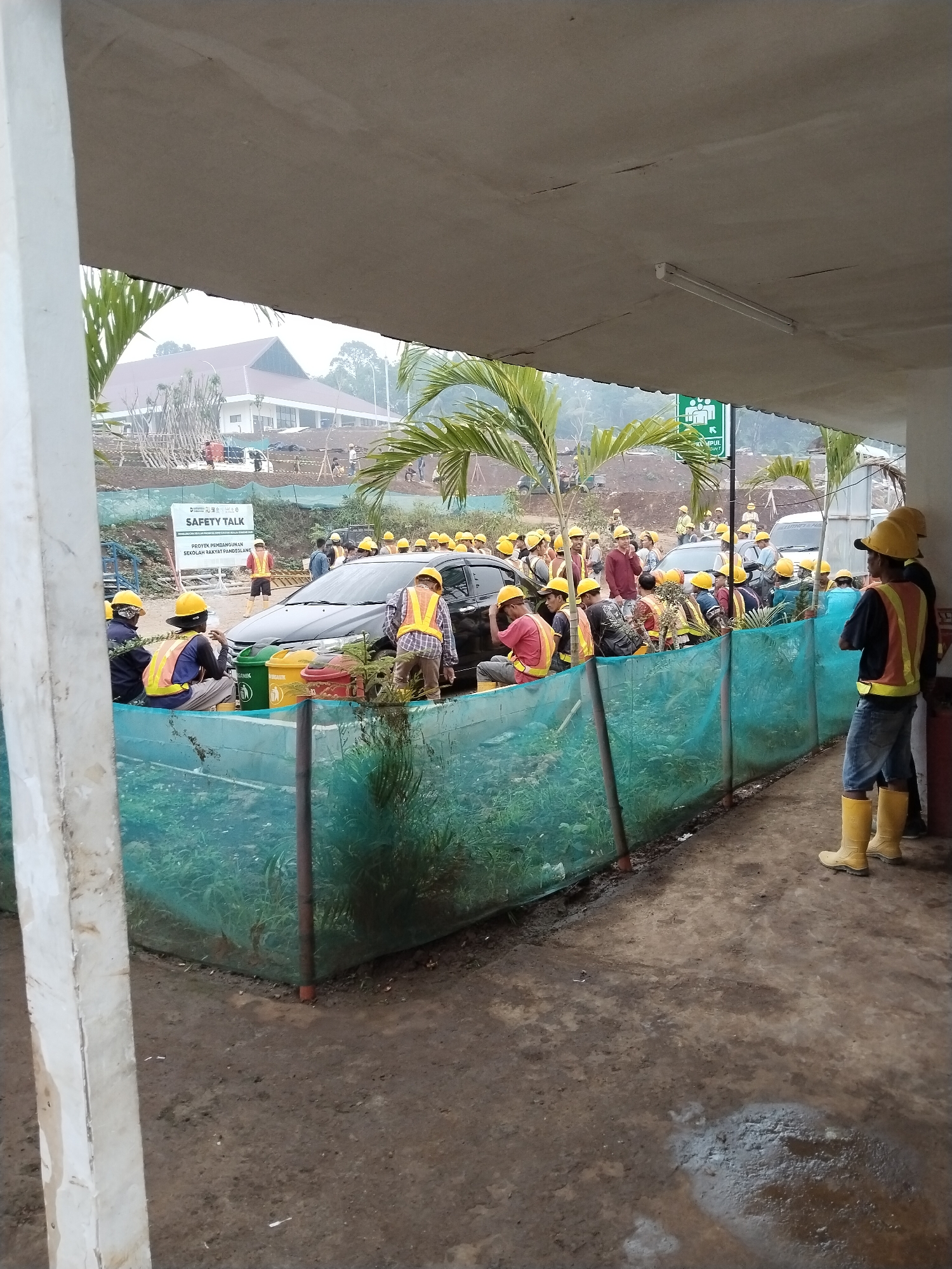 The image depicts a group of construction workers gathered outdoors, likely for a safety talk. A prominent sign in the background reads "SAFETY TALK" in English and also contains Indonesian text that may indicate the location or purpose. Many of the workers are wearing hard hats and safety vests, with some seated and others standing. A black car is partially visible, and several recycling bins are positioned near the workers. The environment appears to be a construction site, with earthworks and some distant buildings visible, suggesting a semi-urban or rural setting. The presence of palm trees indicates a tropical climate. The lighting suggests it is daytime, possibly overcast.