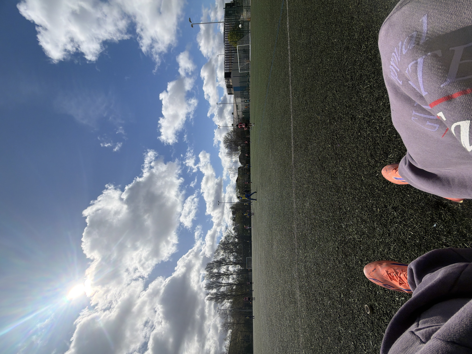 The image shows an artificial turf football pitch on a sunny day in London, United Kingdom. Fluffy clouds fill the blue sky. People wearing sports clothing are playing football on the pitch. A person wearing mauve sweatpants and orange football boots is seated at the edge of the pitch.