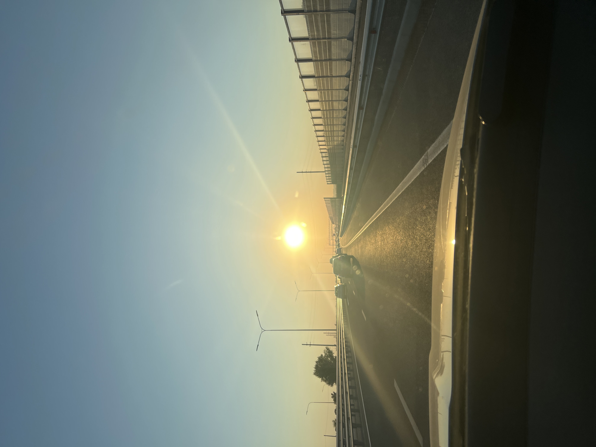 The image depicts a view from inside a vehicle traveling on an elevated highway during sunset. The sun is a bright, glowing orb positioned high in the sky, casting a warm, golden light across the scene. Rays of sunlight flare outwards from the sun, creating a lens flare effect. The sky is a clear, pale blue, transitioning to a warmer yellow hue near the horizon.
The highway itself is multi-lane, with white dividing lines visible. Several vehicles are present on the road, appearing as dark shapes in the distance due to the bright sunlight. A sound barrier, composed of concrete and glass panels, runs along the right side of the highway. Streetlights are also visible along the median of the road. On the left side of the highway, there are some distant trees and utility poles. The foreground of the image shows the hood and windshield of the vehicle from which the photo is taken. The reflection of the sunset is visible on the windshield.
The location is identified as the city of Tretiy Reshayuschiy, Russia. The overall impression is that of a peaceful, serene drive during a beautiful sunset.