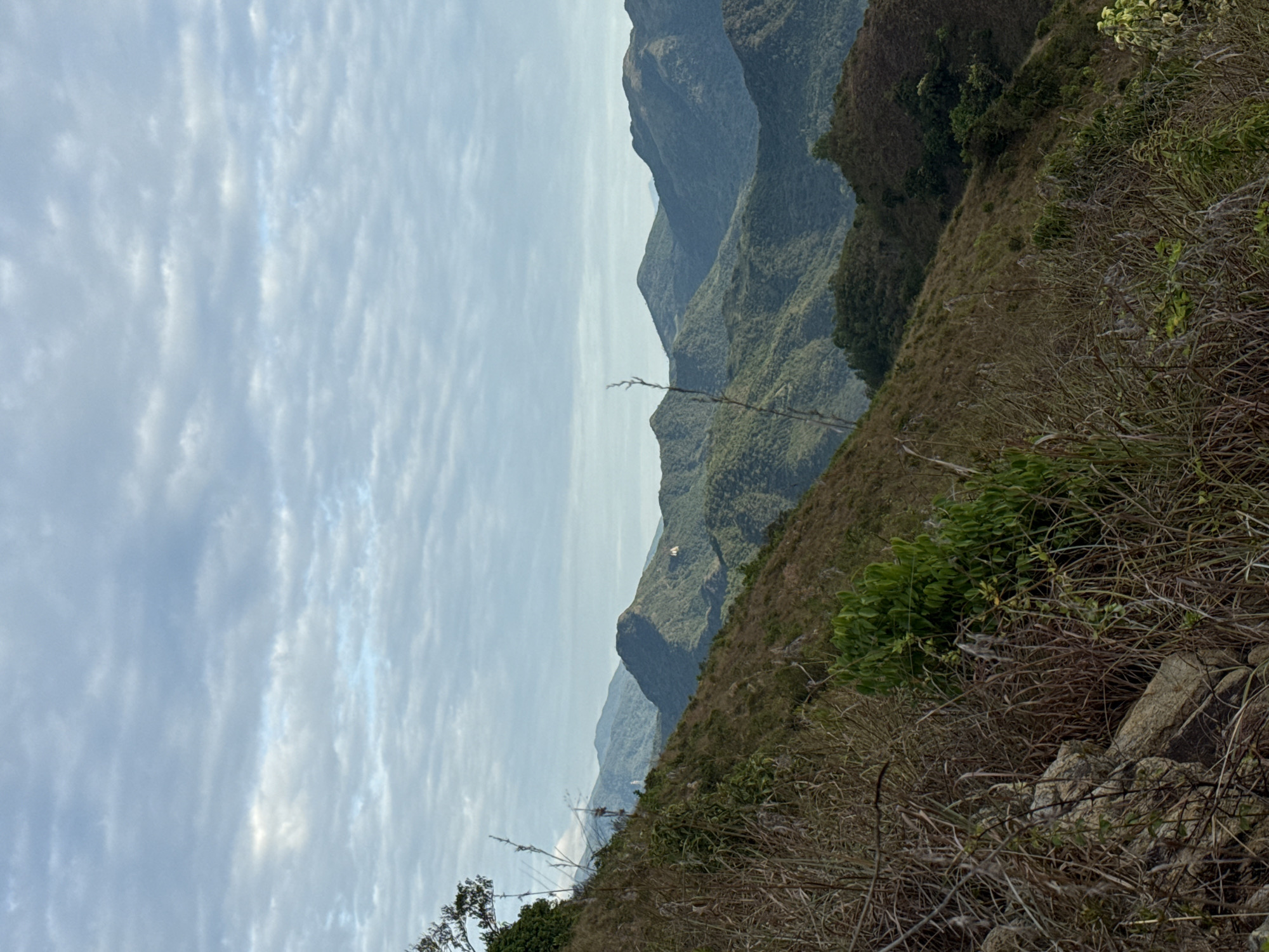 The image depicts a mountainous landscape, likely located in or near Bac Nha Trang Ward, Vietnam. The scene features layers of rolling hills and mountains, partially obscured by a hazy sky filled with thin, streaky clouds. The foreground shows a steep hillside covered in dry grasses and scrub vegetation, with some visible rocks. There are no people visible in the image. The focus is on the natural terrain, suggesting a remote or sparsely populated area.