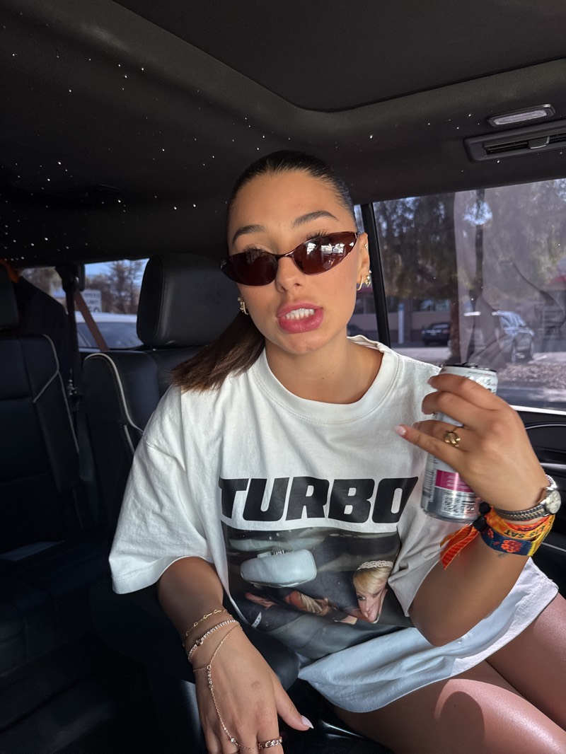 A young woman is seated in the back of a car, looking directly at the camera with her mouth slightly open, revealing her teeth. She has dark hair pulled back into a ponytail and wears dark, slim cat-eye sunglasses. Her skin appears tanned, and she has visible hoop earrings.
She is wearing an oversized white t-shirt featuring a prominent graphic. The word "TURBO" is printed in large black capital letters across the chest, below which is an image showing a person with blonde hair from the chest up, seemingly also inside a vehicle.
Her left wrist displays several delicate, gold-toned bracelets, including one connected by a chain to a ring on her ring finger. Another ring is on her left index finger. In her right hand, she holds a metallic silver can of drink with red and white accents. Her right wrist is adorned with a watch and a colorful fabric wristband, where the letters "SAT" are partially visible.
The car's interior features dark leather seats and a dark headliner embedded with small white lights, resembling a "starlight" effect, typical of luxury vehicles. Bright daylight illuminates the scene from the car's windows, casting shadows within the cabin.