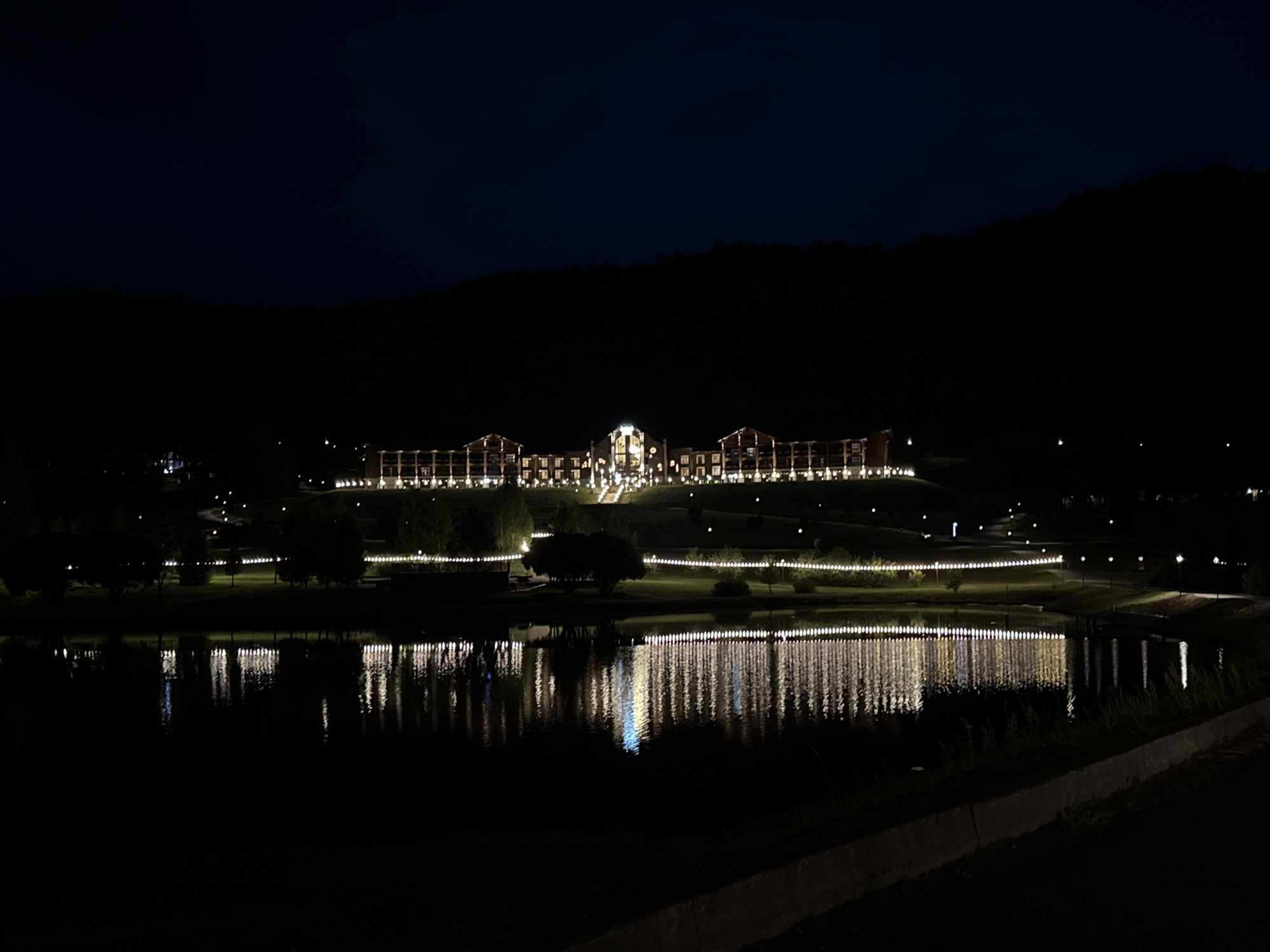 The image shows a large, brightly lit building complex at night, situated on a hill overlooking a body of water. The building has a long, horizontal structure with multiple illuminated windows and architectural lights. String lights also illuminate the landscape leading up to the building. The body of water in the foreground reflects the lights from the building and the surrounding lights. The sky is dark, indicating nighttime. The location is described as Urlu-Aspak, Russia.