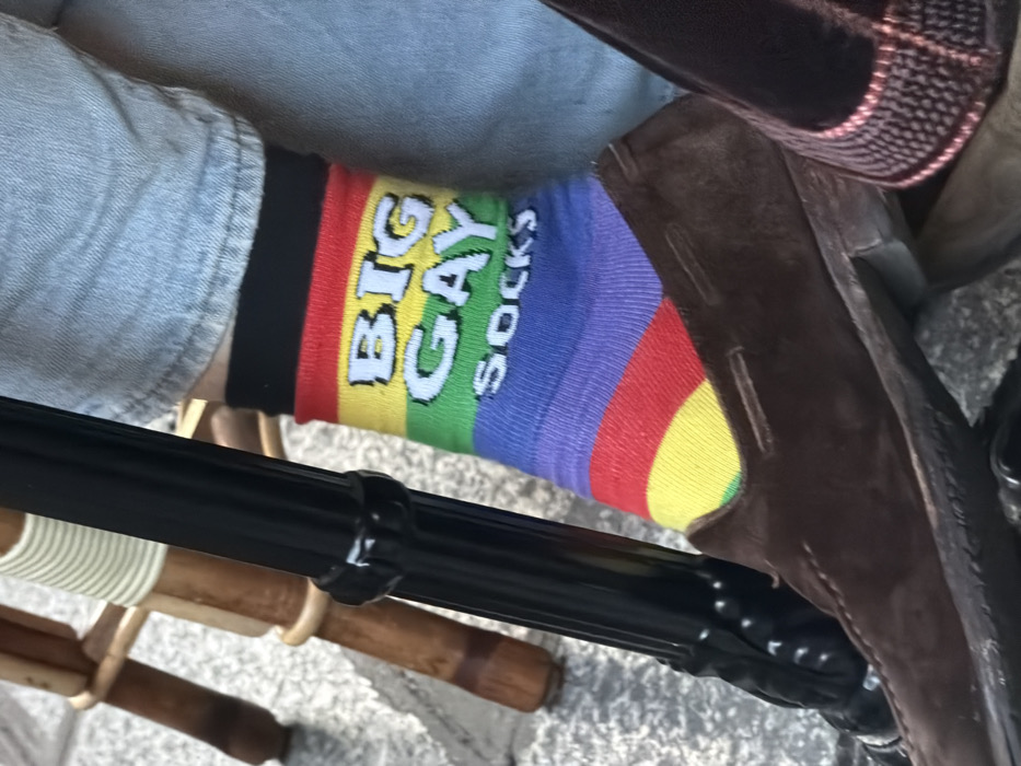 A close-up, low-angle photograph captures the lower left leg and foot of an individual seated in an outdoor cafe in Paris, France. The subject wears light-wash blue denim jeans, cuffed above the ankle. A multi-colored sock, patterned with horizontal stripes in red, orange, yellow, green, blue, and purple, is visible on the foot. White block lettering on the sock reads "BIG GAY" across the yellow and green stripes, with "SOCKS" on the blue stripe beneath it. The foot is encased in a dark brown suede loafer-style shoe. A portion of another dark-colored, textured shoe is visible to the right. A black, fluted metal table leg stands vertically in the left foreground, partially obscuring a light-colored wooden cafe chair leg with wrapped details. The ground is paved with irregular light-grey cobblestones or flagstones.