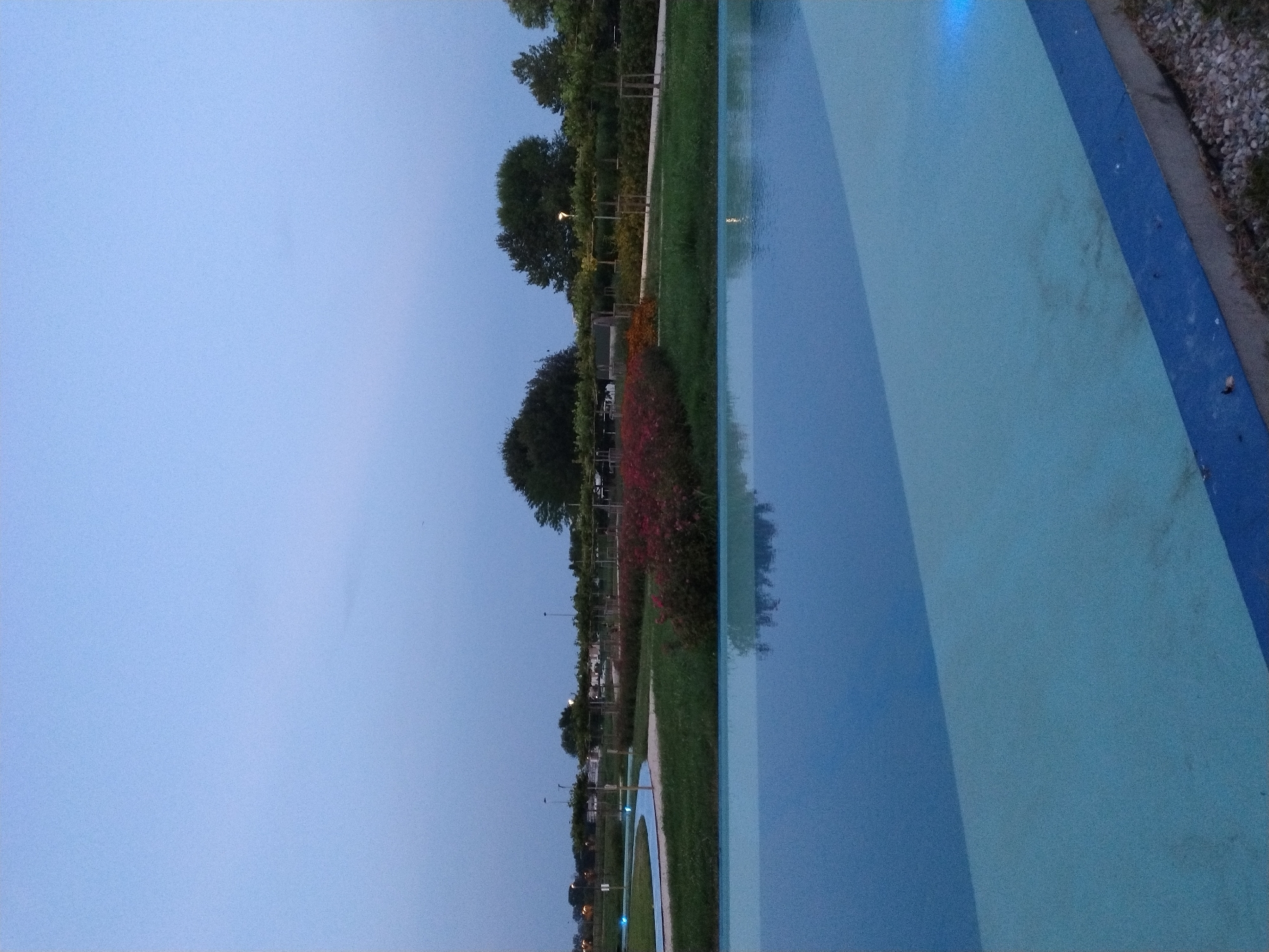 A tranquil, wide-angle shot captures an outdoor public space at dusk. The sky is a soft, hazy blue, transitioning to a slightly darker hue near the horizon. In the foreground, a large, shallow pool of water with a light blue surface reflects the sky and the surrounding environment, creating a serene atmosphere. The water's edge is bordered by a dark blue trim, and beyond that, a strip of gravelly ground and sparse grass.
The middle ground is a lush green lawn interspersed with pathways and meticulously maintained garden beds. A winding, light blue path curves through the grass, hinting at a recreational area. Flowerbeds bursting with vibrant red and pink blooms add a splash of color to the scene, contrasting with the soft greens of the grass and the dark foliage of trees and shrubs. Several young trees with neatly pruned branches stand at intervals, adding structure to the landscape. Streetlights, subtly illuminated, dot the background, suggesting a public park or recreational facility. The background features a line of trees and a glimpse of distant buildings and infrastructure.
The scene appears to be located in the Parco Archeologico di San Zaccaria, a public park in Caorle, Italy, known for its archaeological finds and recreational facilities. The overall impression is one of peacefulness and order, with the park bathed in the soft light of twilight. No people or animals are visible in the image.