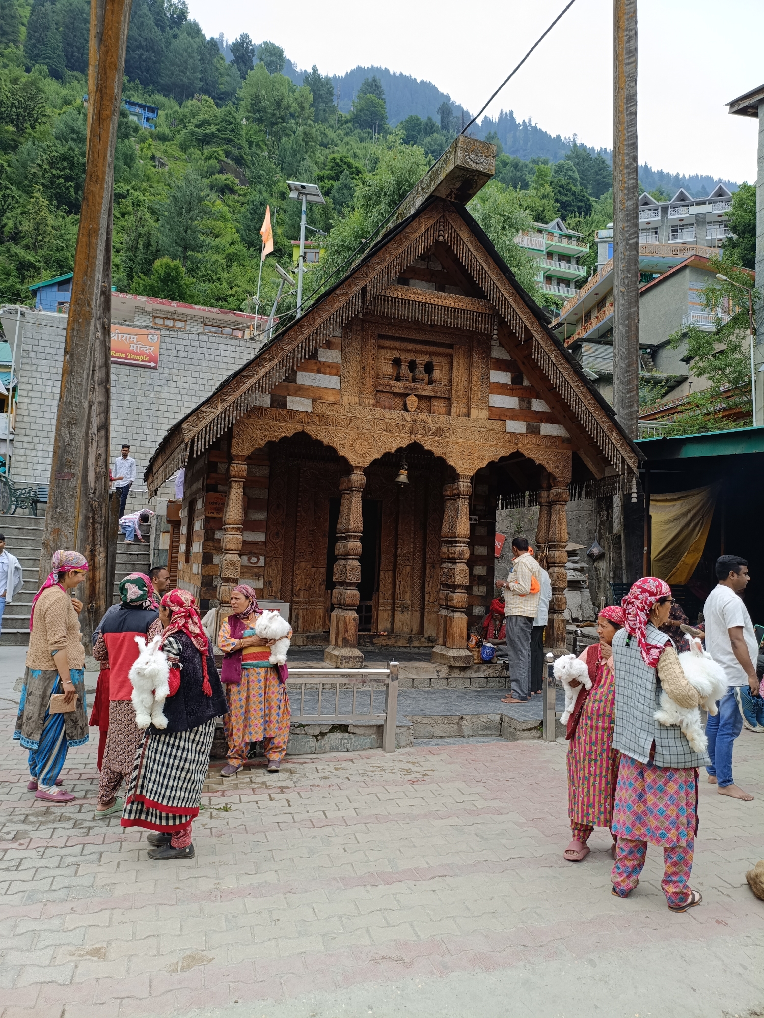 The image depicts the entrance to the Ram Temple in Vashisht, India, a town nestled in the Himalayan mountains. The temple, constructed of intricately carved wood, features a traditional Himachali architectural style with a sloped roof adorned with decorative wooden detailing. Several individuals, predominantly women dressed in traditional Indian attire with headscarves, are gathered in the foreground. Some are holding fluffy white animals, possibly rabbits or sheep.
The ground in front of the temple is paved with interlocking bricks. Behind the temple, a brick building with a sign indicating "Ram Temple" is visible. The background is dominated by a lush green mountainside covered with dense forest and scattered buildings, characteristic of a mountain town. A few men are also present, including one standing on the temple steps and another in the middle ground observing the scene. The overall atmosphere suggests a lively cultural and religious site.