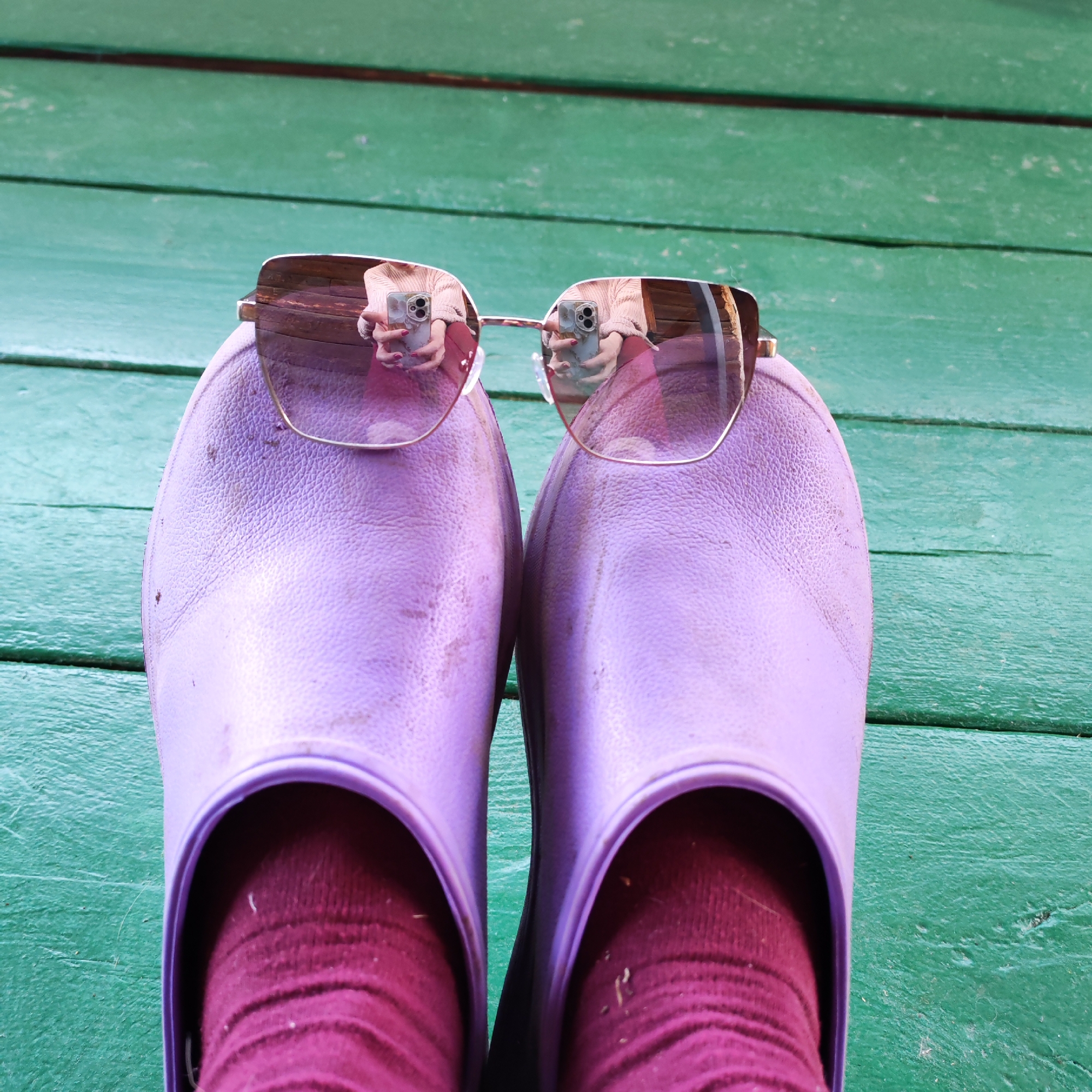 The image shows a first-person view of a pair of lilac-colored slip-on shoes worn with maroon socks, resting on a green wooden surface. A pair of sunglasses with a gold frame and light brown lenses is perched on the shoes. The reflection in the lenses shows a person holding a smartphone, likely taking the photograph. The location may be in the city of Chaikovsky, Russia but there are no elements of the picture that confirms this.