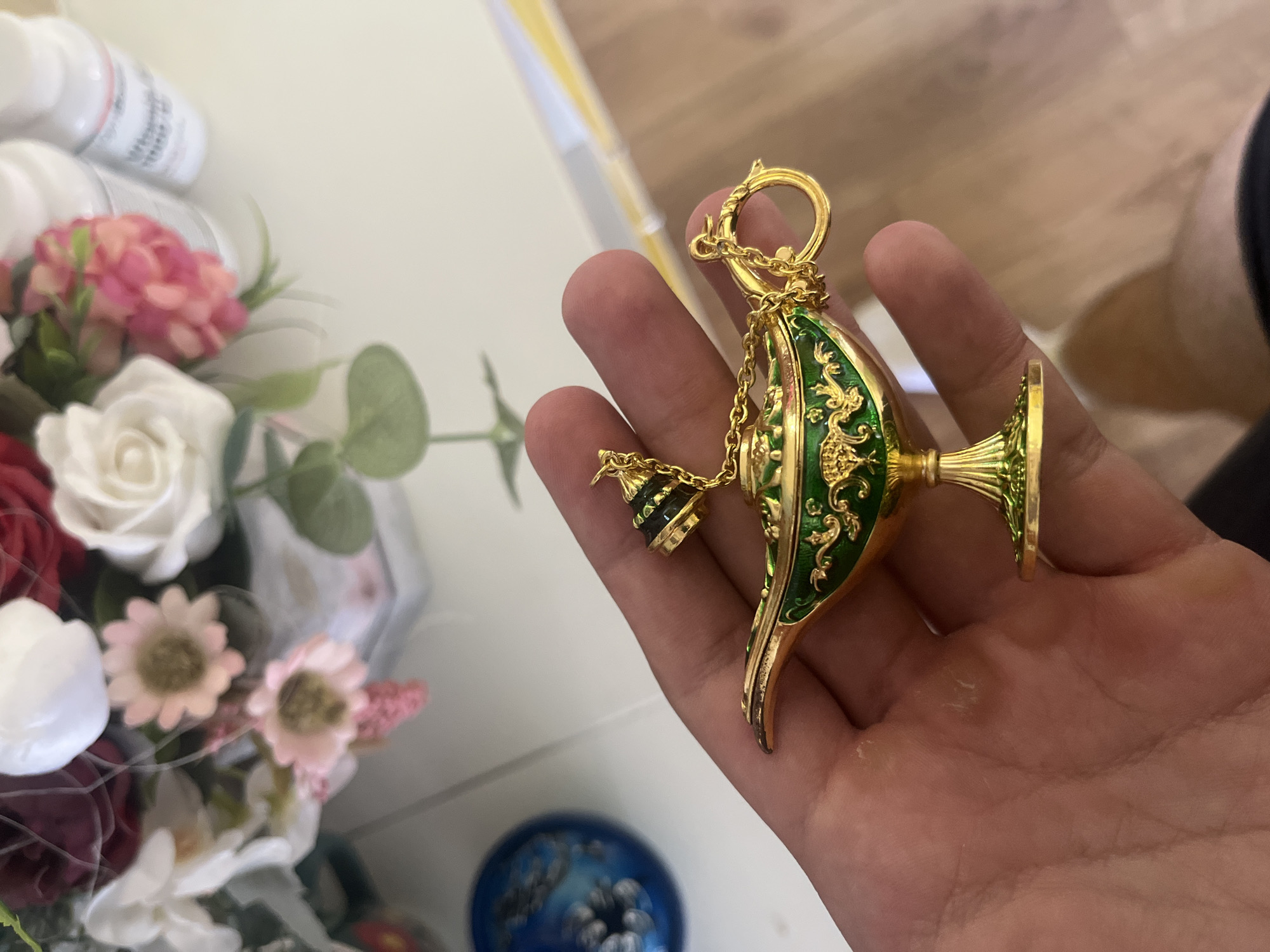 A hand is holding a decorative Aladdin's lamp keychain. The keychain is gold and green, with intricate scrollwork on the body of the lamp. A small chain connects the lamp to its lid, which is also adorned with green and black detailing. The background of the image is blurred, but appears to be a tabletop with flowers and bottles. The scene is indoors, with natural light. No specific location in Pokrovnik, Bulgaria is discernible from the image.