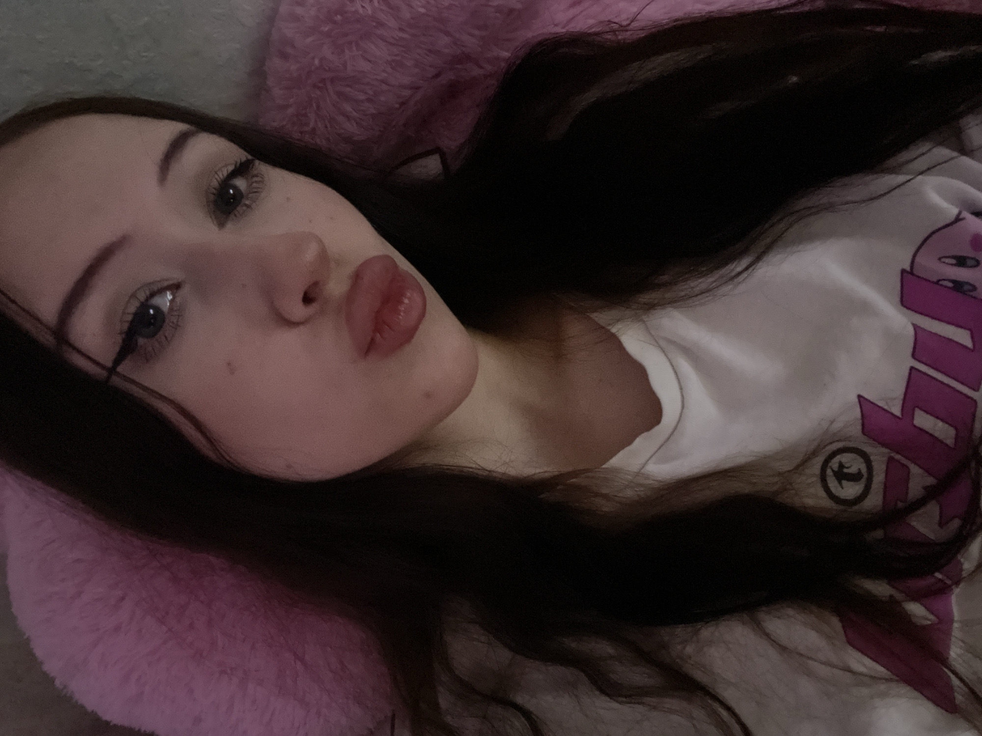 The image is a close-up, self-taken photograph of a young person lying down, possibly in bed. The subject has long, dark hair and is wearing a white t-shirt with a large, stylized purple graphic on it. The graphic appears to be text-based with some abstract elements and a circular emblem. The person's face is turned towards the camera, with their lips puckered in a kiss. Their eyes are open and looking at the camera, and they have winged eyeliner. The background is soft and out of focus, with a pink, fuzzy texture, suggesting a pillow or blanket. The lighting is dim, indicative of indoor lighting, possibly at night. The overall mood is playful and intimate. The image does not contain any overt cues to Votkinsk, Russia, beyond the provided location context.