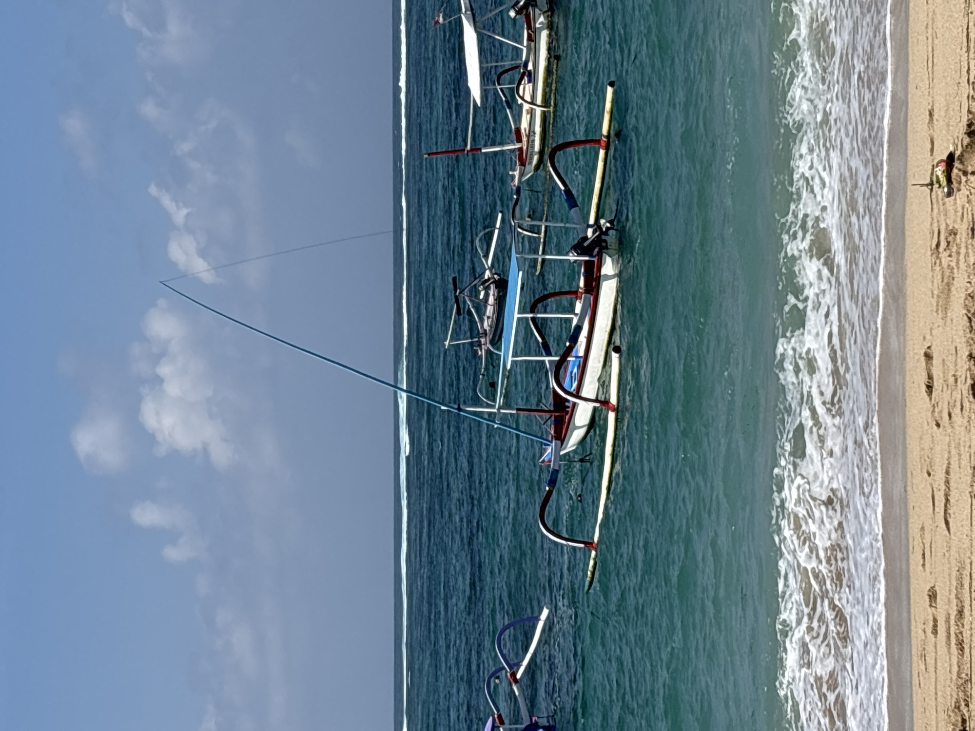 A bright, clear day with a few scattered clouds illuminates a serene ocean scene in Kuta Selatan, Indonesia. The turquoise waters are calm, with gentle waves lapping at a sandy beach in the foreground. Several traditional Indonesian outrigger boats, known as jukungs, are anchored offshore. The boats, painted in white with accents of red, blue, and purple, are equipped with fishing rods or poles. In the distance, a thin white line of breaking waves marks the horizon. The scene is peaceful and idyllic, with the boats resting on the water and the beach awaiting visitors.