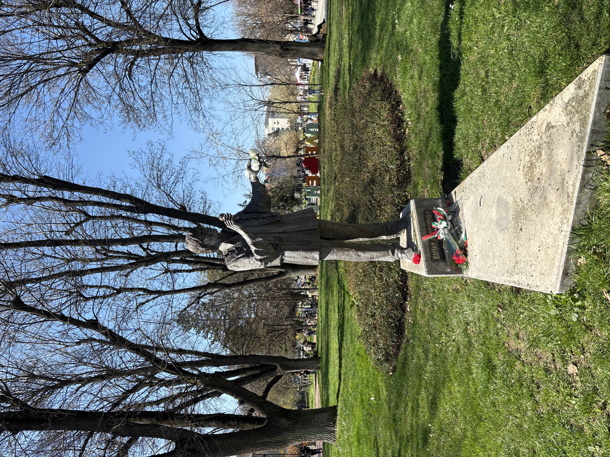 Here's a description of the image:
The image depicts a statue in what appears to be a park setting in Pleven, Bulgaria. A concrete sculpture of a person stands on a concrete plinth. The statue holds a bouquet of white flowers. On the plinth, there are several bouquets of red flowers with ribbons. Grass surrounds the statue, and a low shrubbery feature is behind it. Bare trees are prominent in the scene, lining the park in the background. In the distance, there are buildings and people strolling along a path. The sky is blue and mostly clear. The location is likely a public park or memorial site, given the presence of the statue and floral tributes.