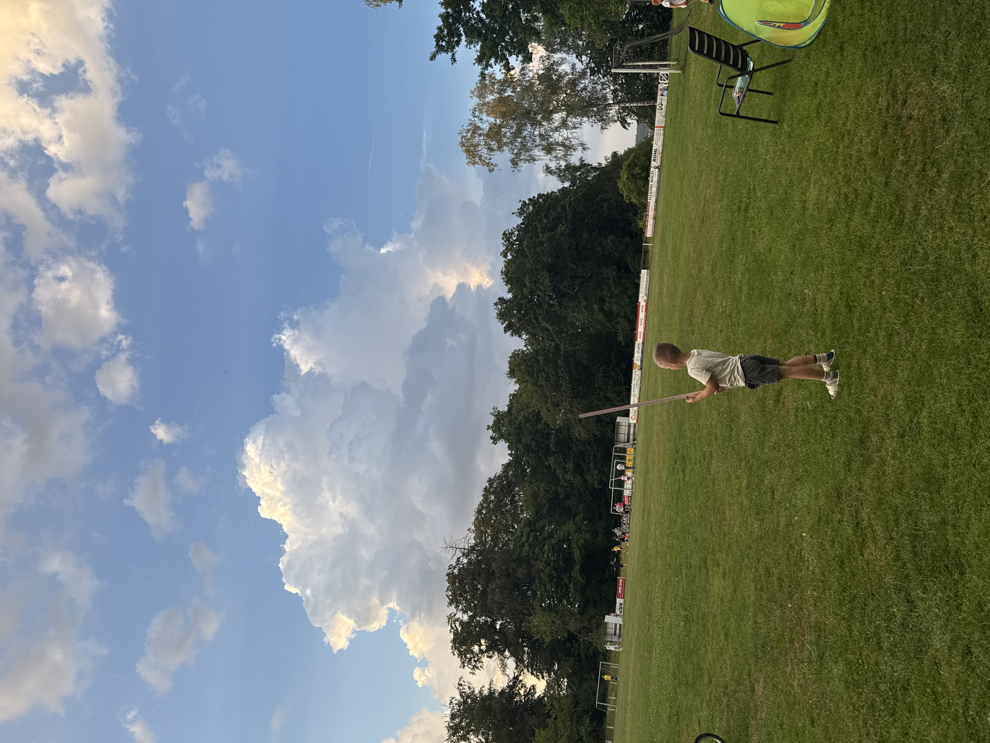 A young boy stands on a green grassy field, holding a thin, long object, possibly a stick or a pole, vertically in front of him. He is wearing a light-colored t-shirt and dark shorts, with white socks and shoes. In the background, a soccer game is in progress on the same field. Several players in uniforms are visible, along with soccer goals. A line of trees forms a backdrop beyond the field, and above them, a dramatic sky with large, white, illuminated clouds stretches across the frame. To the right of the boy, a black chair and a yellow tent-like structure are present. The overall setting suggests an outdoor recreational area, potentially a park or sports field, in Lumda, Germany.