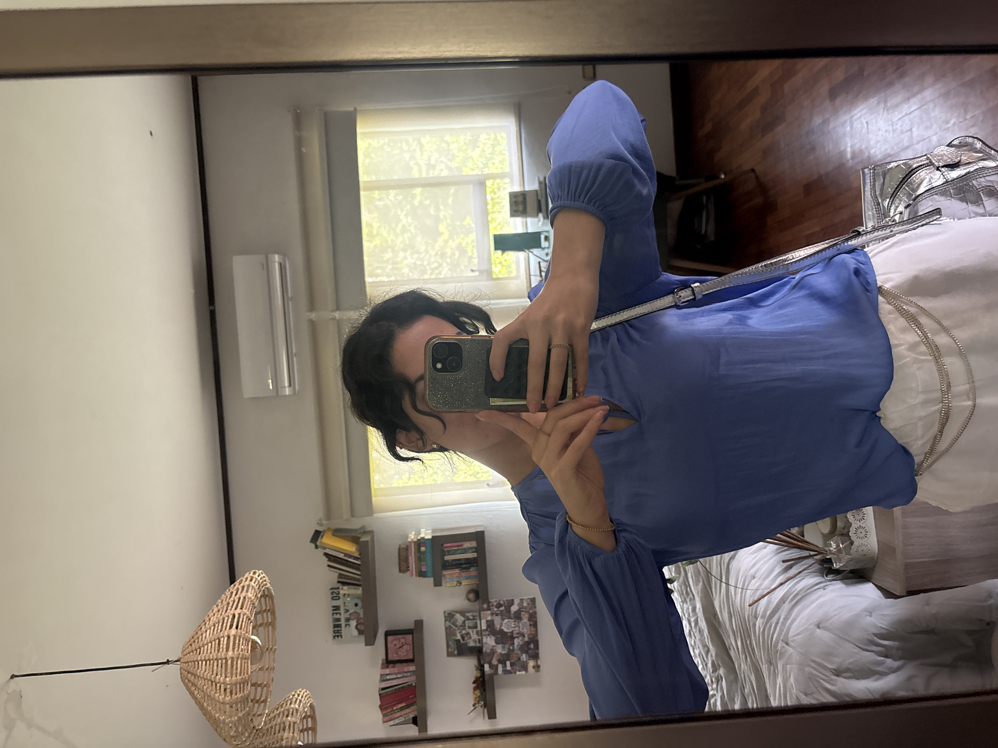 A woman is taking a mirror selfie in a room. The woman is wearing a blue blouse and white pants. There is a silver purse to the right of her. Behind the woman is a window with natural light streaming in. To the left of the window, there is a wall-mounted air conditioner. Underneath the window, there is a bookshelf with books and pictures on it. In the bottom left corner of the image is a wicker light fixture. The floor is wooden. The city and country are not discernible from the image.