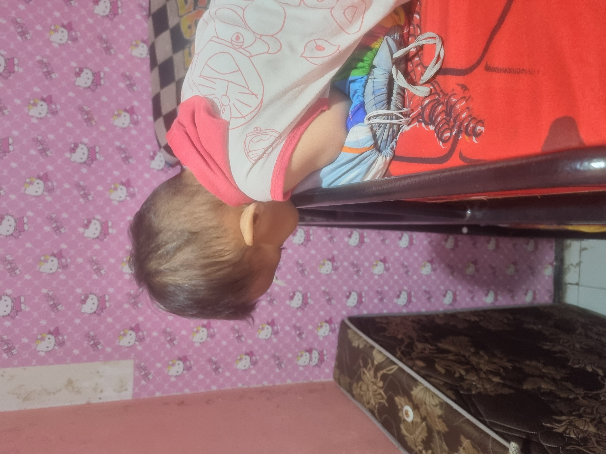 A young child is lying on their side on a red surface, appearing to be a bed or mattress. The child is wearing a white sleeveless shirt with pink trim and a pink hood, featuring a cartoon character illustration in pink outline. The child's face is turned away from the camera, and their head is resting on colorful fabric. The background consists of pink wallpaper with a repeating pattern of cartoon cat characters. To the left of the child, a brown mattress with a floral pattern is visible. The scene appears to be indoors, possibly a bedroom. The image does not provide specific details to pinpoint a location within Probolinggo, Indonesia.