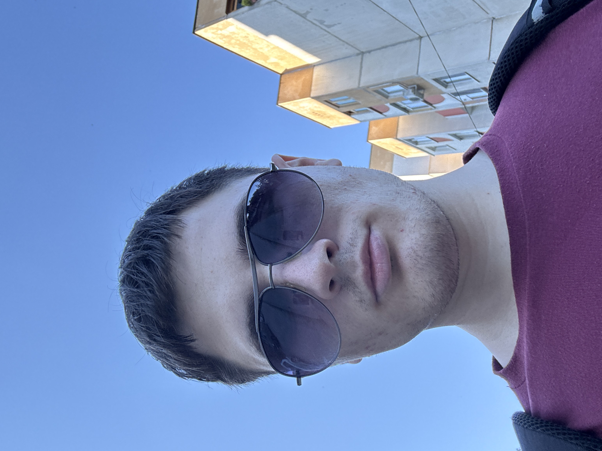 A low-angle, medium shot captures a young Caucasian male facing forward, wearing aviator sunglasses and a maroon crew-neck t-shirt. His dark hair is short and styled casually. In the background, a bright blue sky dominates, with the upper portions of several apartment buildings visible on the right. These buildings exhibit a layered architectural style with balconies and windows, suggesting an urban setting. The lighting indicates it is daytime, with the sun casting a slight yellow hue on the buildings. The image appears to be a selfie taken outdoors in Stara Zagora, Bulgaria.