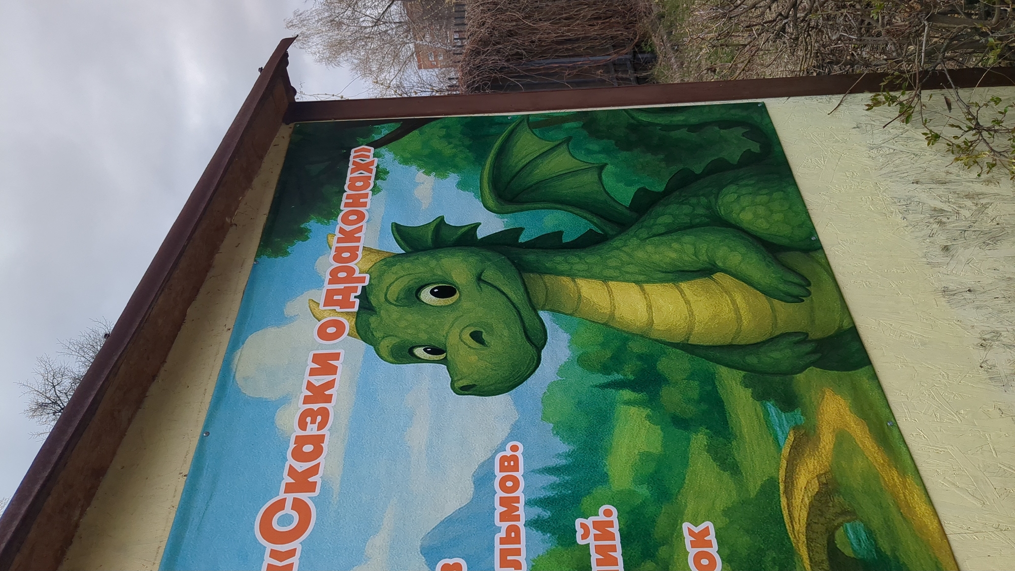 An outdoor sign, likely a billboard or large informational panel, is depicted in an angled shot against an overcast sky. The main feature of the sign is a vibrant, cartoon-style illustration of a friendly green dragon. The dragon has large, dark eyes, small horns, and substantial wings, with a lighter yellow-green belly. Its body is scaled, and its expression appears gentle and slightly curious.
The dragon is set against a backdrop of a bright blue sky with soft white clouds on the left, transitioning into a lush green landscape or forest on the right. The artwork suggests a fantastical or children's story theme.
Visible Russian text on the sign, rendered in prominent orange letters, reads "Сказки о Драконах," which translates to "Tales about Dragons." Below this title, several partial words are visible: "В," "...льмов.", "...ий.", and "ОК". These likely indicate authors' names or publishing details for the dragon tales.
The sign itself has a brown, possibly metal, frame along its top and left edge, and the main image is affixed to a lighter, yellowish board. Surrounding the sign are bare tree branches, some with very small, emerging green leaves, suggesting early spring. In the distant background, a building and more trees can be seen. The overall setting is an outdoor, possibly urban or park area in Ulyanovsk, Russia. The sky is uniformly cloudy, indicating an overcast daytime.
