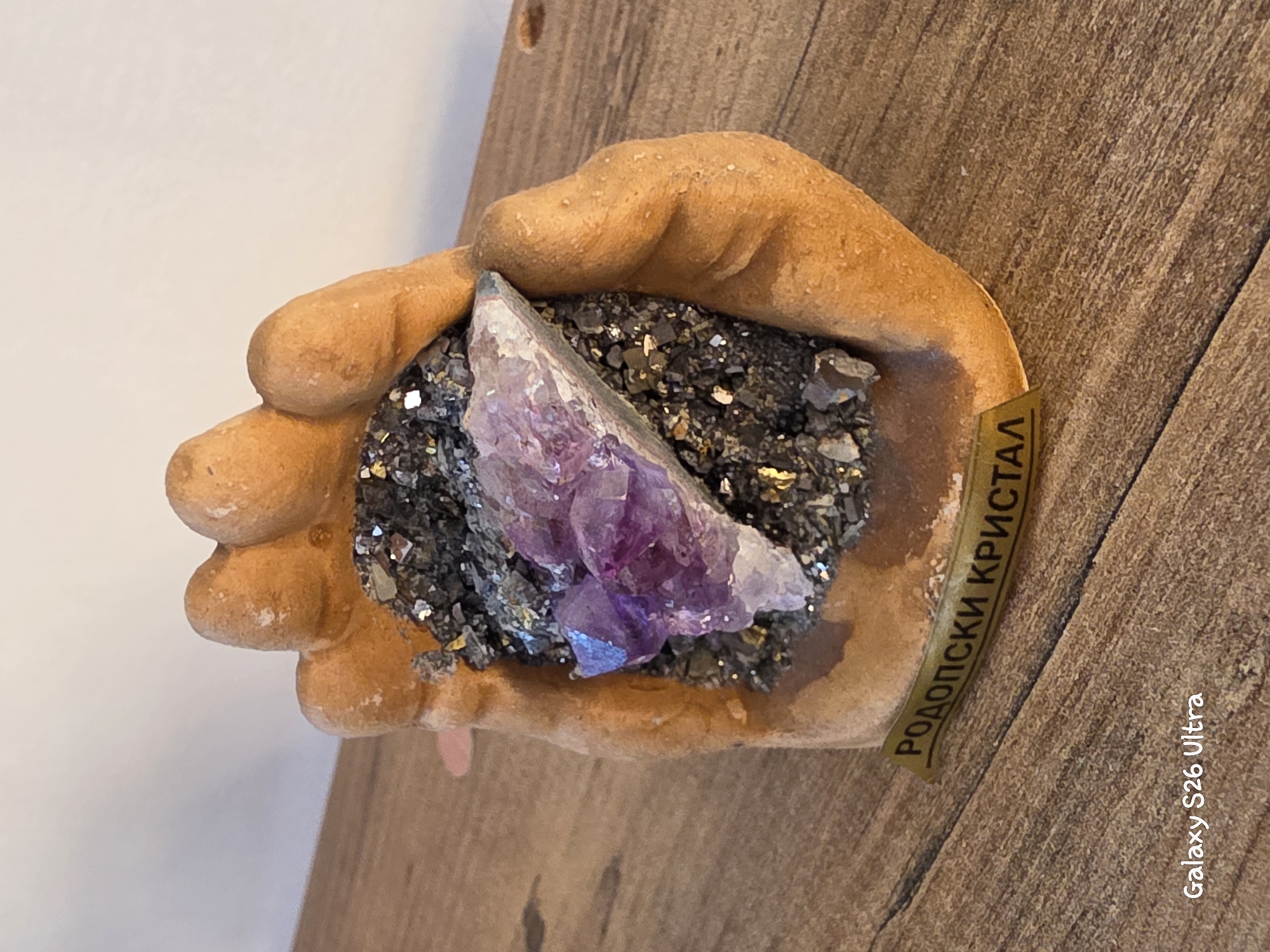 The image displays a close-up shot of a decorative item, possibly a souvenir, featuring a cluster of amethyst crystals embedded in dark, metallic-looking rock. This cluster is held within a sculpted, terracotta-colored hand. A golden label with Cyrillic text, "РОДОПСКИ КРИСТАЛ" (Rodopski Kristal), is affixed to the base of the hand. The item rests on a wooden surface, likely a table or shelf, which exhibits a visible wood grain. The background is a plain, light-colored wall. The bottom left corner of the image displays a watermark: "Galaxy S26 Ultra". There are no people or animals present in the scene. The object appears to be an artistic representation related to the Rhodope Mountains region of Bulgaria, which is geographically near Plovdiv.