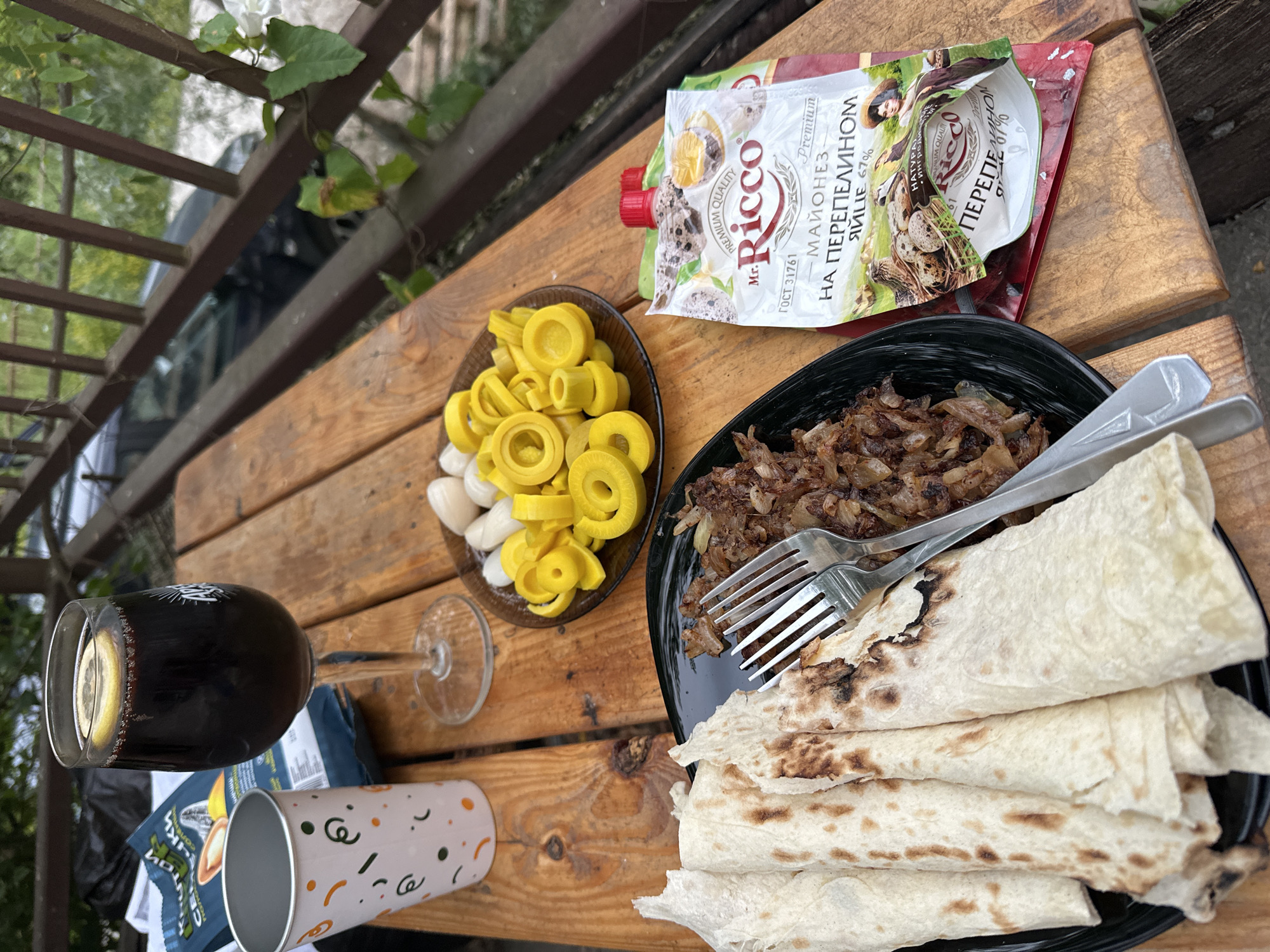 This media file shows a close-up, overhead view of a meal laid out on a rustic wooden table, likely outdoors. The scene suggests a casual meal, possibly a picnic or a relaxed outdoor gathering.
The subjects include a plate of food, a glass of dark liquid with a lemon slice, a packet of mayonnaise, a small bowl of yellow pickled items and some white objects, and a decorative paper cup. The main dish appears to be some kind of filling, possibly meat or vegetables, served with flatbread or lavash. A fork rests on the plate, indicating the meal is ready to be eaten.
The packet of mayonnaise has visible text in Russian, identifying it as "Mr. Ricco Premium Mayonnaise on Quail Egg 67%". The yellow items in the bowl are likely sliced pickled vegetables, and the white items could be pickled garlic or small onions. The drink in the glass is dark, possibly wine or juice. A partially visible bag of what looks like sunflower seeds is also present. The overall setting is a wooden table, with a blurry background suggesting an outdoor environment with some greenery and a railing, hinting at a patio or balcony.
There are no people visible in the image, but the presence of a prepared meal implies an activity or event of dining. The lighting suggests it could be daytime, with natural light illuminating the scene. The overall mood appears relaxed and informal.