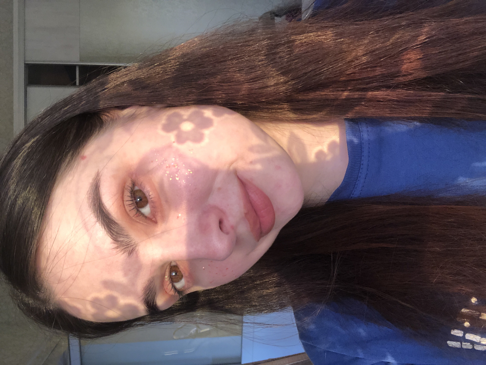 A young woman with brown hair and brown eyes is depicted in a close-up photograph. She is wearing a blue t-shirt and has flower-shaped shadows cast on her face. Her skin appears to have glitter on it. The background shows a window and part of an interior wall. It is unclear how this is related to Noviy, Russia.