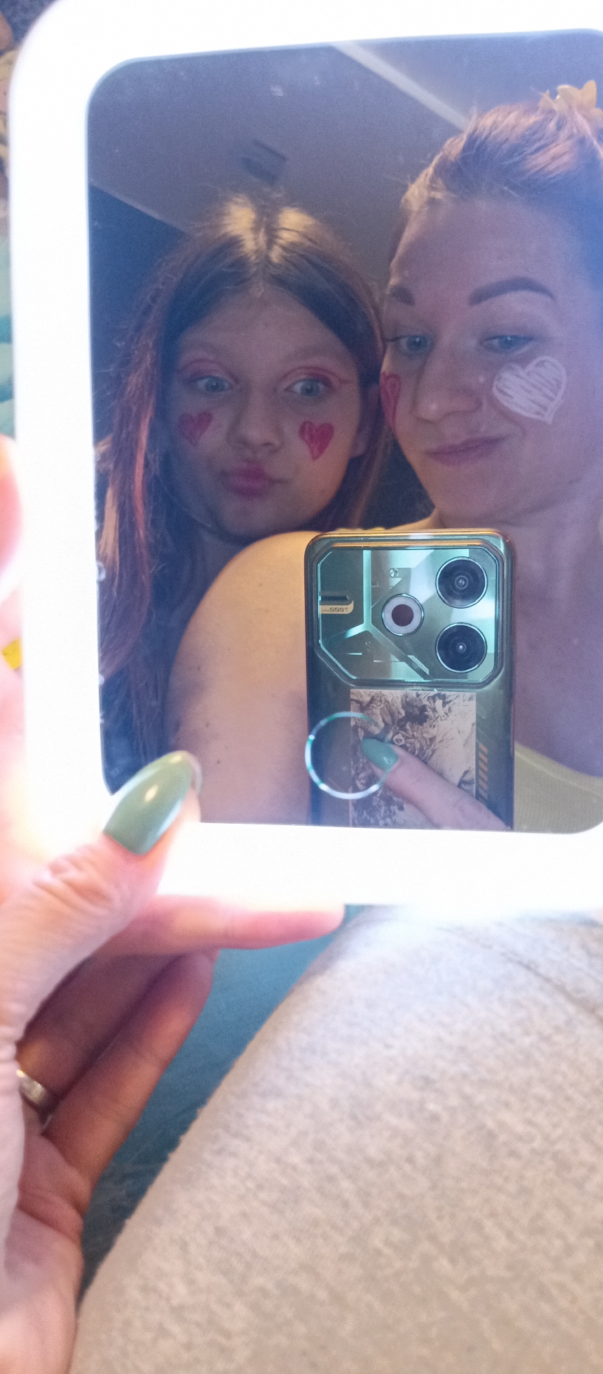 A selfie captures two people in a mirror, one a younger person, and the other an adult. Both individuals have heart designs drawn on their faces. The younger person wears red hearts near their eyes, while the adult has a white heart on their cheek. A hand holding a smartphone is visible in the foreground. The phone has a decorative case and a ring grip. The background appears to be an interior setting. The location in the city of Unknown, Unknown, is unknown.