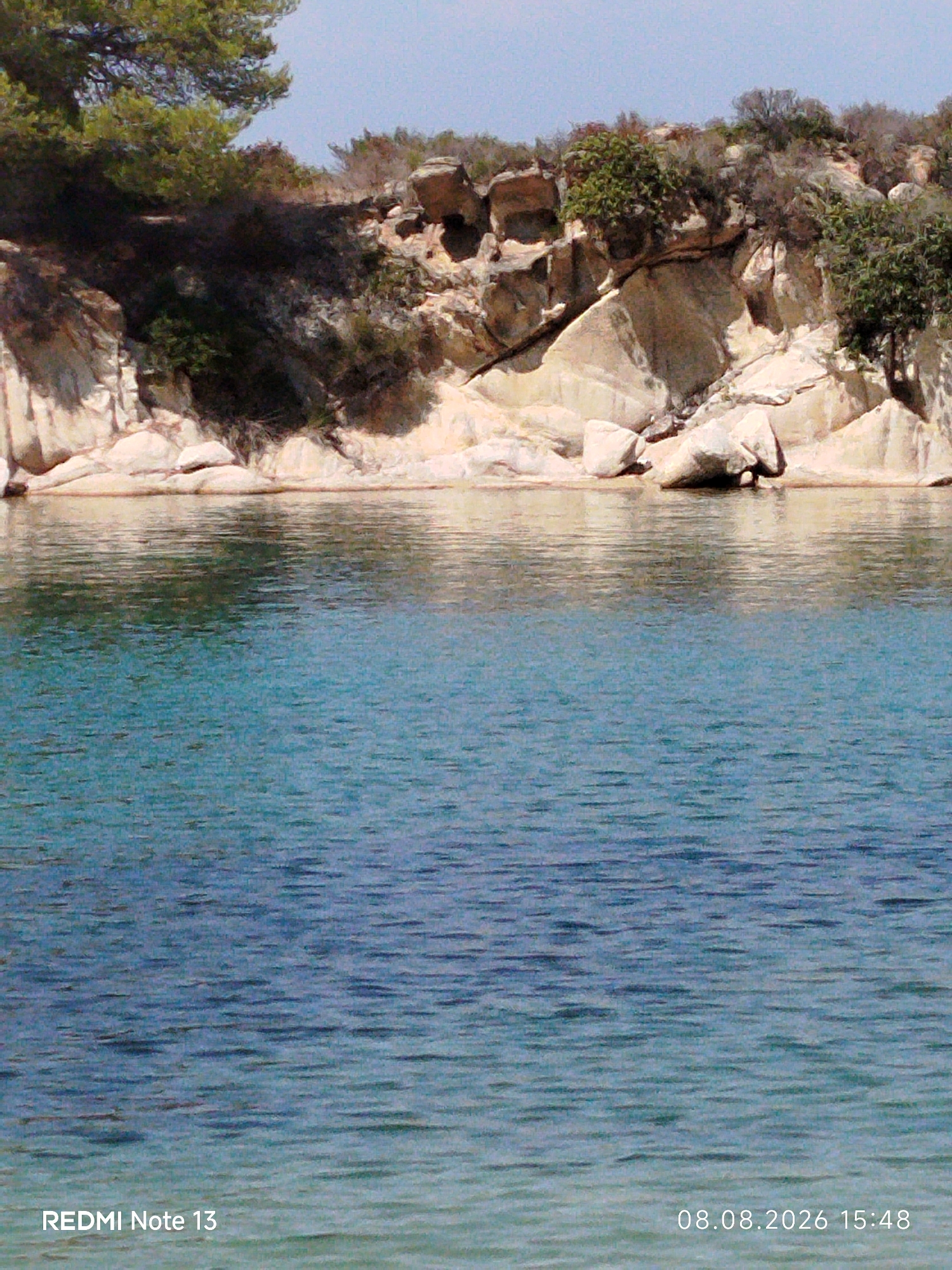 A waterfront view shows a rocky coastline with sparse vegetation against a clear blue sky. The foreground is dominated by calm, turquoise-colored water with subtle ripples. In the background, a shoreline is characterized by large, light-colored rocks and patches of green foliage, suggesting a natural coastal environment. The bottom corners of the image display overlaid text: "REDMI Note 13" on the left and "08.08.2026 15:48" on the right. The scene appears to be a tranquil natural setting, likely a beach or cove within Sithonia Municipal Unit, Greece. There are no people or discernible actions depicted.