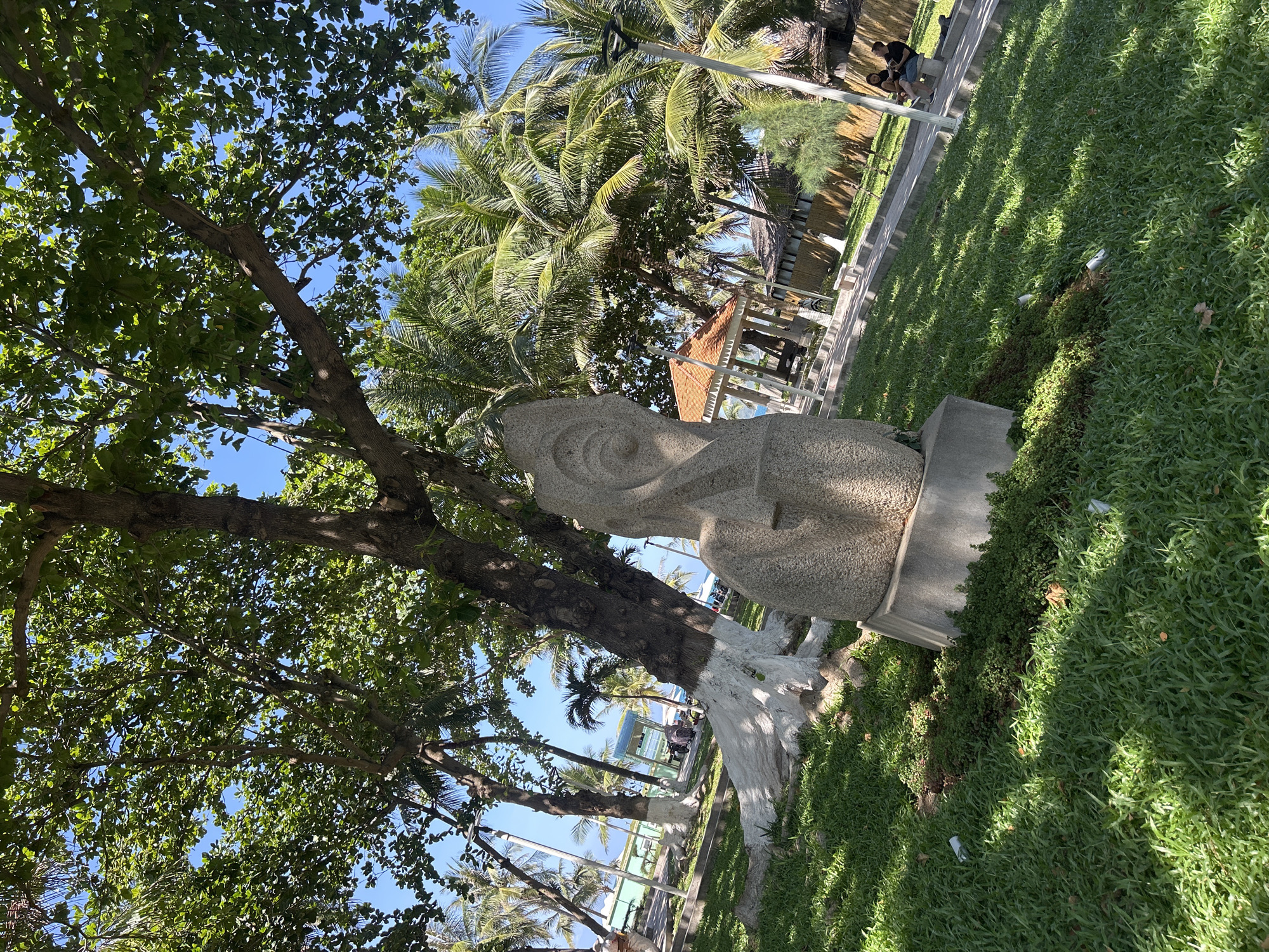 The image captures a landscape in Nha Trang Ward, Vietnam, featuring a weathered, light-colored stone sculpture situated on a concrete base amidst a grassy area. The sculpture is partially obscured by the branches and foliage of a large tree, the trunk of which is painted white at the base. Palm trees and other vegetation are visible in the background. A structure with a thatched roof is also partially visible, suggesting a recreational or public space. In the upper part of the frame, figures of people can be seen sitting along a pathway. The scene is bathed in sunlight.