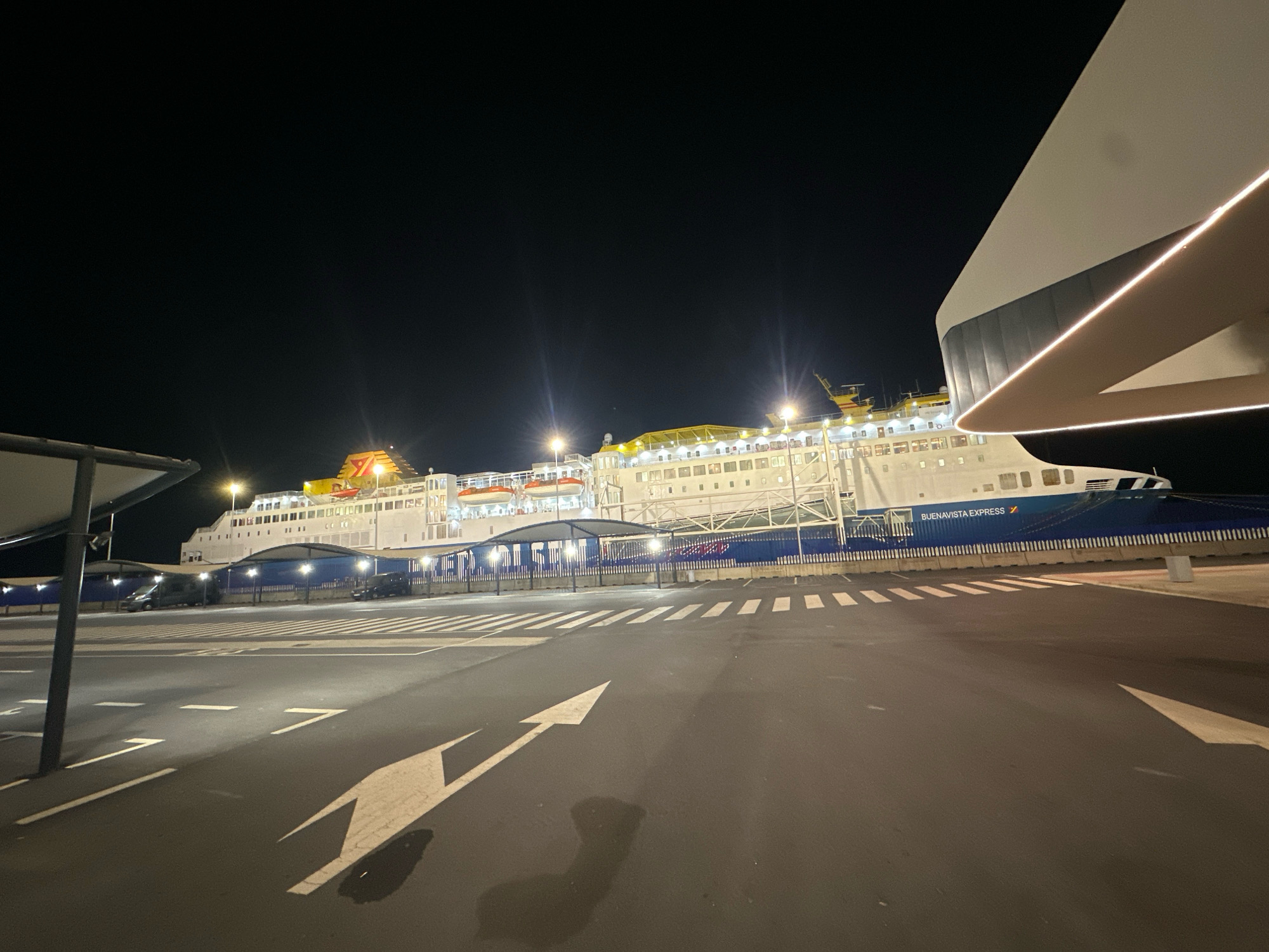 The image shows a large white ferry, the "BUENAVISTA EXPRESS," docked at night in the port of Palos de la Frontera, Spain. The ferry is illuminated by exterior lights, highlighting its numerous windows, lifeboats, and yellow accents on its upper decks. In the foreground, a dark asphalt surface with white directional arrows and pedestrian crossing markings suggests a parking lot or waiting area. A modern, curved white structure with integrated lighting is partially visible on the right side of the frame, possibly part of the terminal building. The background is a dark night sky. A few dark-colored vehicles are parked under covered shelters along the dockside, in front of the ferry. The overall scene depicts a port facility at night, with the ferry prominently displayed.