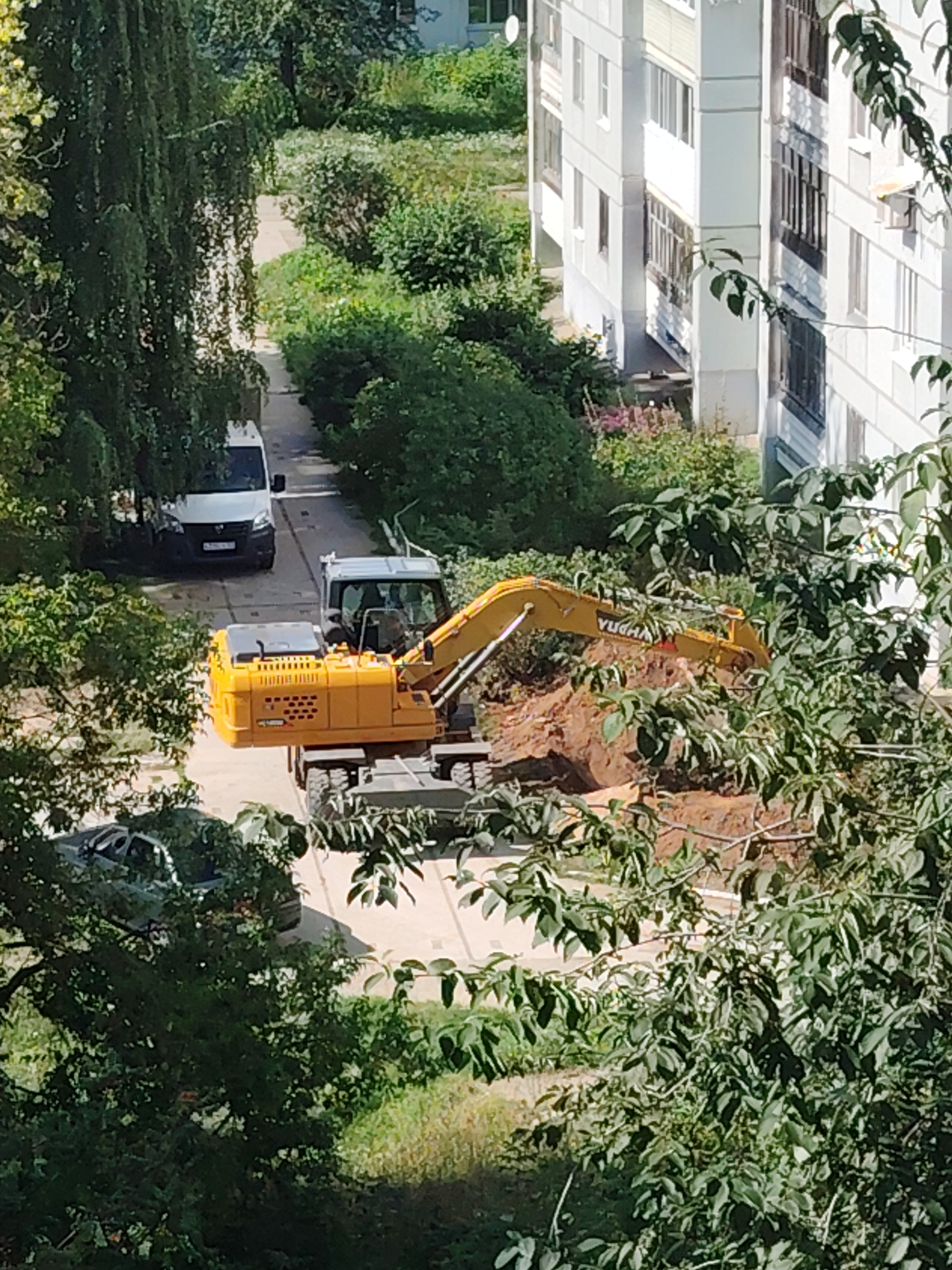 The image shows an exterior scene in the city of Chaikovsky, Russia, likely a residential area given the presence of an apartment building. A bright yellow excavator is positioned in a dirt excavation, with its boom arm extended. Nearby, a white van is parked on a paved surface, and a dark-colored car is partially visible. Lush green trees and bushes dominate the foreground and surround the construction site. The apartment building, appearing to be several stories high, is white with dark-framed windows and balconies. The overall impression is of ongoing construction or repair work within a somewhat overgrown urban environment.