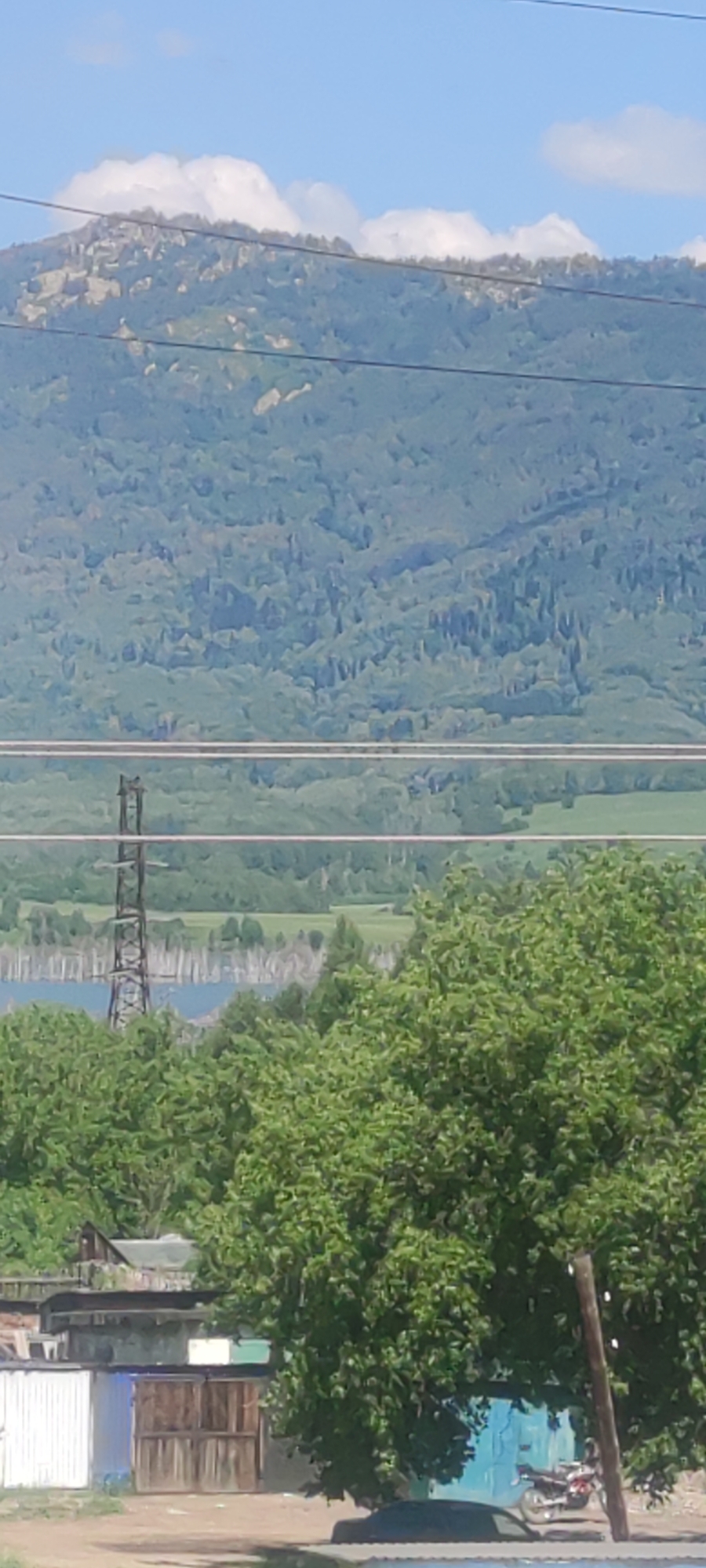 The image shows a landscape with mountains in the background, partially covered by green trees. A power line tower is visible in the middle ground. In the foreground, there are some buildings, including what appear to be sheds or garages, with a motorcycle parked near one of them. The overall scene suggests a rural or semi-rural setting. The location is specified as the city of Ridder, Kazakhstan.