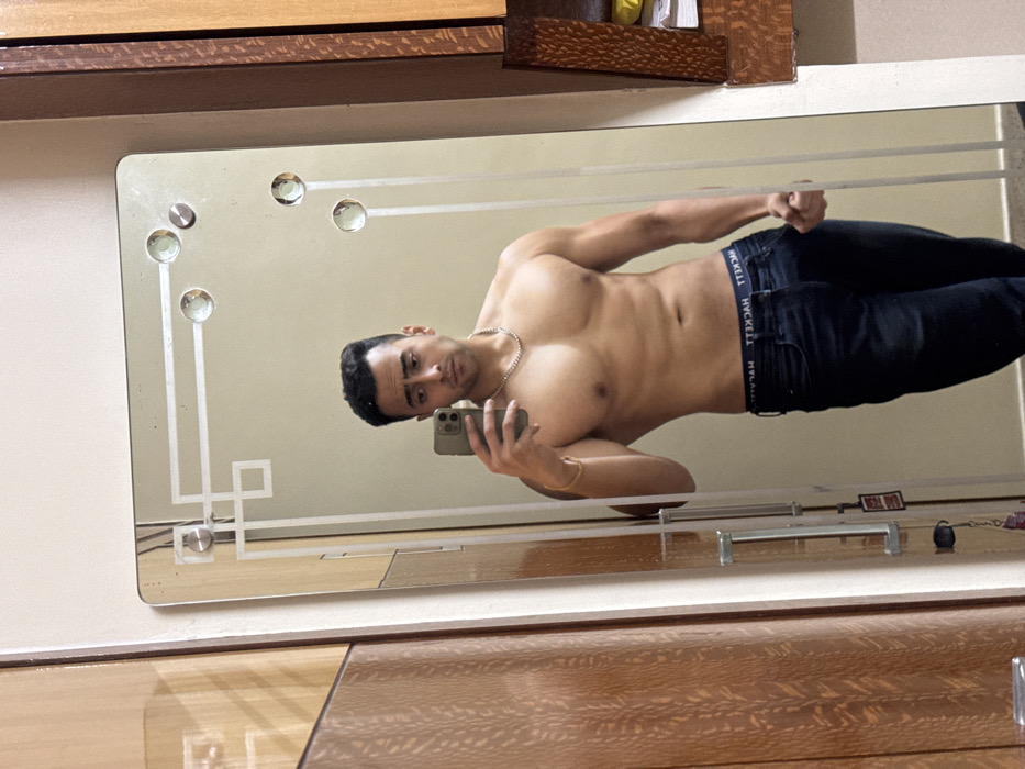 The image displays a mirror selfie of a muscular man indoors, likely in a home setting in Kolkata, India. The image is rotated 90 degrees counter-clockwise, presenting the subject horizontally across the frame.
The central subject is a young adult male with dark hair and a serious expression, looking directly into the camera. He is shirtless, revealing a toned physique, and is wearing dark-wash jeans. A white chain necklace and a gold-colored bracelet are visible accessories. His right hand holds a smartphone, presumably an iPhone, with which he is taking the picture reflected in the mirror. His left hand is clenched into a fist at his side.
The waistband of his underwear, peeking above his jeans, clearly displays the brand name "HYCKELL" in white text against a dark background.
The mirror itself is rectangular and features decorative elements, including several circular, faceted studs and etched geometric lines in its border, particularly in the top-left and bottom-left (as viewed in the rotated image) corners. The background reflected in the mirror appears to be a plain, light-colored wall.
Below the mirror, there is polished wooden furniture, possibly a dresser or chest of drawers, which reflects the upper part of the room and the mirror itself. The handles of the furniture are visible. The overall setting suggests a private room such as a bedroom or dressing area. The time of day cannot be determined, but the lighting appears even and indoor.