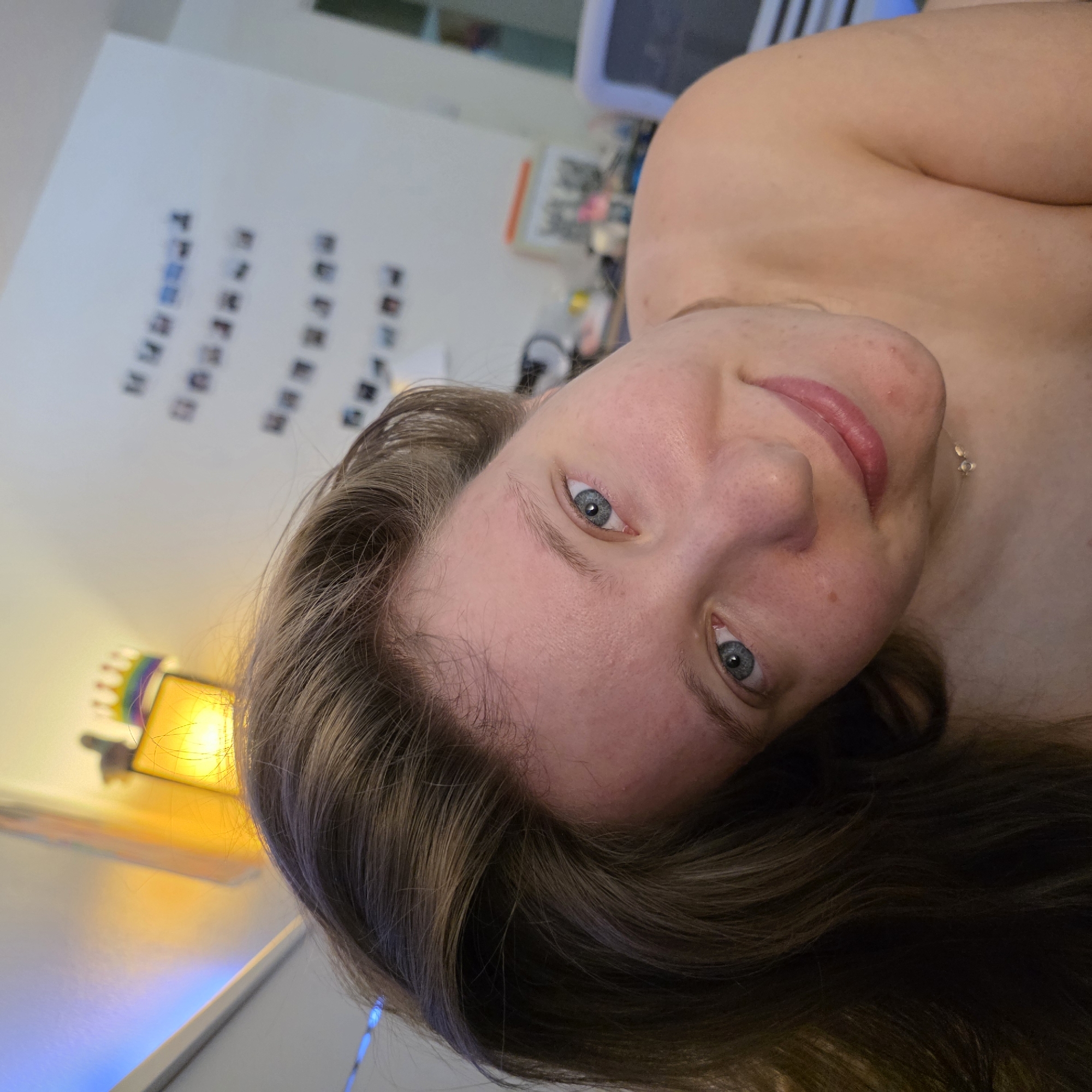 This is a close-up, eye-level, selfie-style shot of a person with fair skin and light brown hair, positioned in a domestic indoor setting. The subject's face is angled towards the camera, and their hair is falling forward around their head. Their eyes are open and looking forward, and they have a slight smile. A faint necklace with two small pendants is visible around their neck. The background is softly blurred, showing a wall with several small framed pictures or photos displayed in rows, a yellow lamp with a rainbow-colored crown-like decoration, and what appears to be a desk or shelf with various objects including what could be a computer monitor. There is also a blue light casting a subtle glow on the upper left portion of the image. The image does not contain any specific landmarks or clues that would indicate a location in Portland, United States of America.