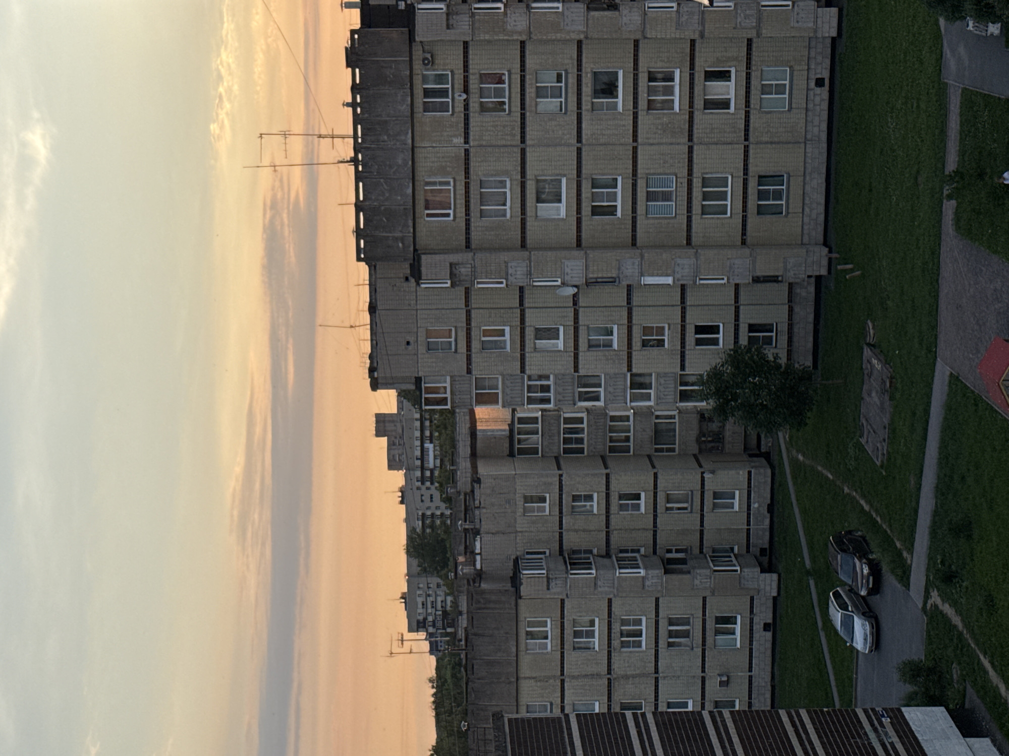 The image depicts a view of apartment buildings in the city of Kirishi, Russia, at dusk. The sky is a gradient of orange and yellow, with some wispy clouds. In the foreground, two apartment buildings, constructed from concrete blocks with visible brickwork, are prominent. These buildings have multiple floors with numerous windows and balconies. Several cars are parked on a paved area in front of the buildings, surrounded by a grassy lawn. In the background, other apartment buildings and structures are visible, some with antennas on their roofs. The overall scene conveys a residential urban environment during twilight.