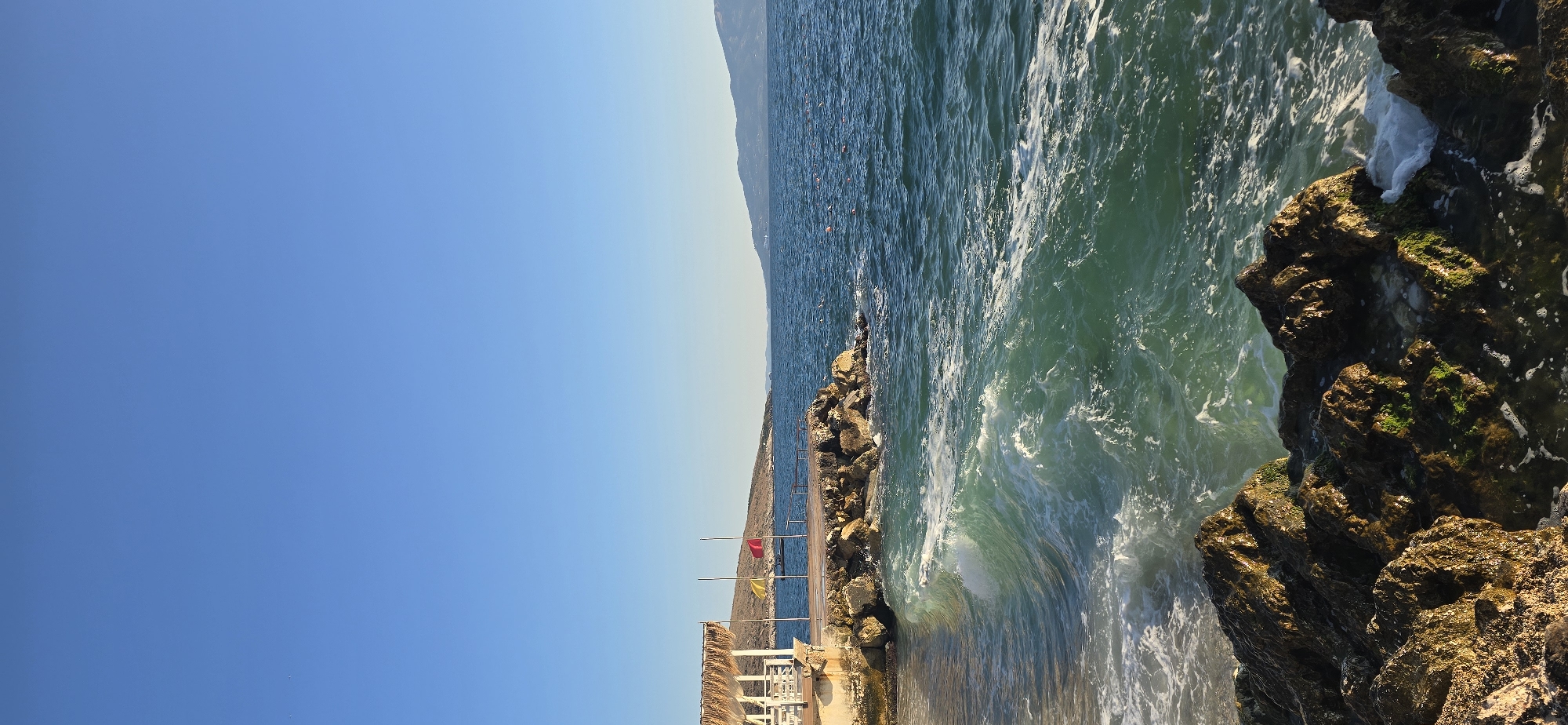 The image captures a serene coastal scene in Saranda, Albania, featuring a clear blue sky and the tranquil waters of the Ionian Sea. In the foreground, rough, dark rocks are partially submerged in the foamy, turquoise water, indicating the edge of a small beach. A gentle wave is breaking against these rocks, creating white foam and spray.
To the left, a small, open-air structure with a thatched roof and white pillars stands on a stone foundation, suggesting a beachside cafe or seating area. Adjacent to this structure is a small wooden pier extending into the sea. Two flagpoles stand on the pier, one with a yellow flag and another with a red flag. A small boat is visible in the water behind the pier.
The middle ground is dominated by the expanse of the blue sea, with a few buoys scattered on its surface. In the distance, the coastline of Saranda is visible, characterized by rolling hills that transition into the mountainous landscape of Albania. The overall atmosphere is peaceful and idyllic, typical of a sunny day in a coastal city. There are no people visible in the image.