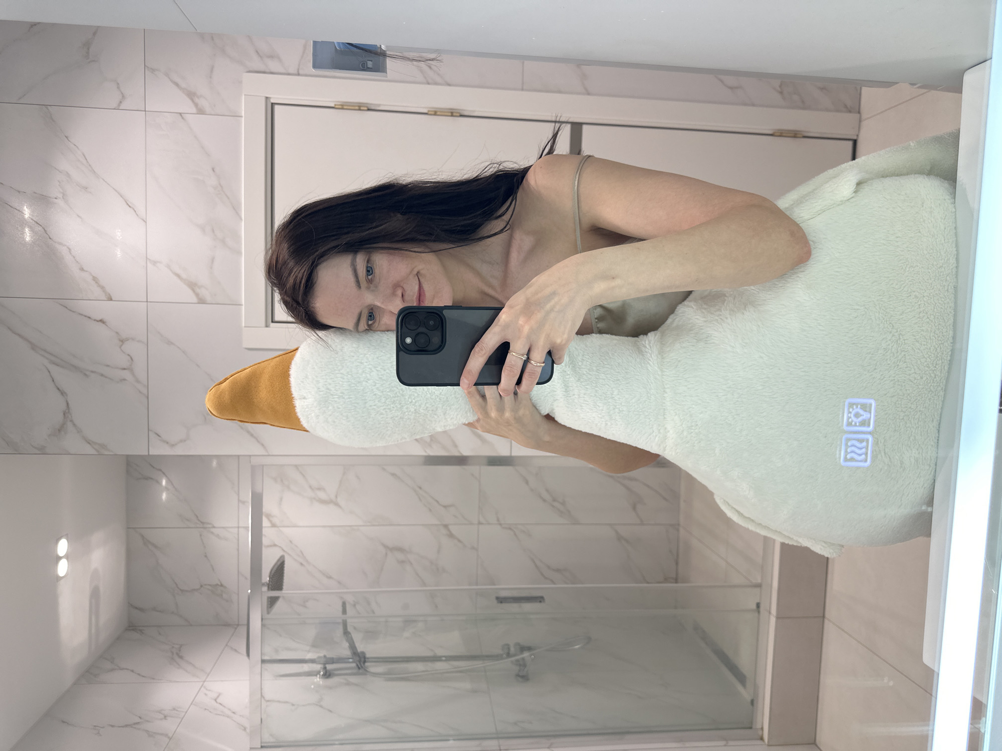 A woman with dark hair is taking a selfie in a bathroom mirror. She is holding a black smartphone and has her cheek resting against a plush, white and orange goose. The bathroom features white marble-patterned tiles on the walls and floor. A glass-enclosed shower is visible in the background. A lighted mirror is to the right. There is no information in the image to place it within the city of Прикамский, Russia.