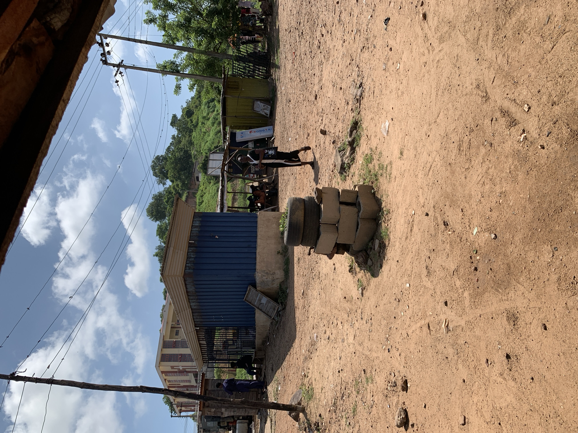 The image depicts an outdoor scene in a dusty, open area that appears to be a market or commercial hub in Lokoja, Nigeria. The ground is covered in reddish-brown dirt with scattered debris and tire tracks. In the foreground, a stack of cinder blocks topped with several tires serves as a visual anchor.
Several structures are visible. To the left, a blue corrugated metal building with a fenced-off entrance is present, and to its left, a partially visible building with a light-colored facade and red accents is situated on a slight incline. Further to the right, a smaller, yellowish corrugated metal structure with an open front and a canopy is seen, with a black metal fence behind it.
People are present in the scene, though their activities are not fully discernible. A person in dark clothing is walking away from the camera towards the right side of the image. Two individuals are near the blue building, one standing and one seated. In the background, several people are gathered near the yellowish structure. Electrical poles and wires are strung across the sky, indicating the presence of utilities. The sky is partly cloudy with a bright blue expanse. The overall impression is one of a functional, perhaps informal, commercial area under a bright, sunny day.