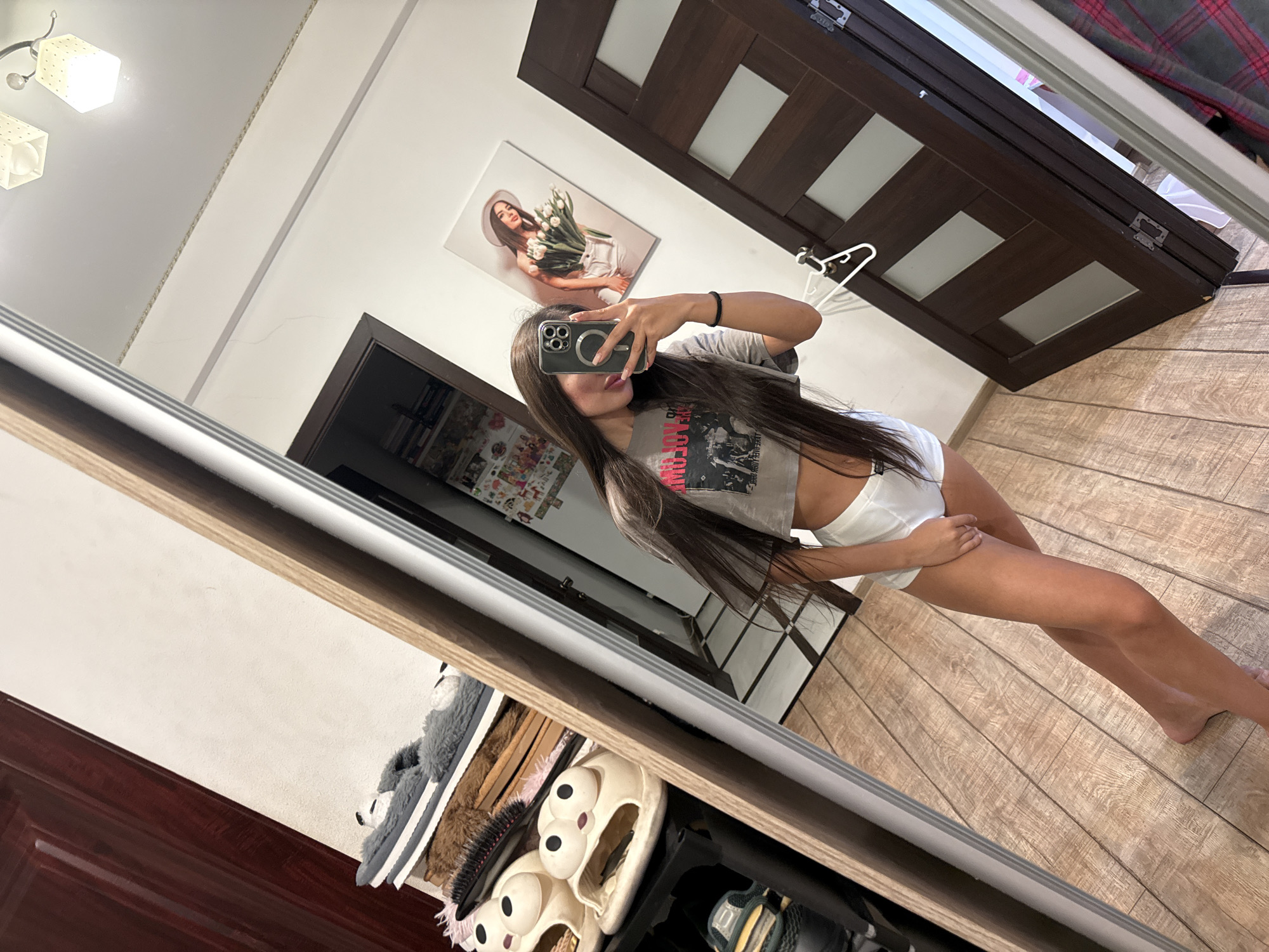A young woman is taking a mirror selfie in a room. She is wearing a grey crop top and white shorts. Her hair is long and dark brown. She is holding a smartphone in her right hand and is partially obscuring her face with it. In the background, there is a shoe rack with several pairs of slippers, a dark wooden door, and a framed picture on the wall of a woman holding tulips. The floor is made of light-colored wooden planks. The image appears to be taken indoors, possibly in a residential setting. There is no specific information in the image to determine the location in the city of Chișinău, Moldova.