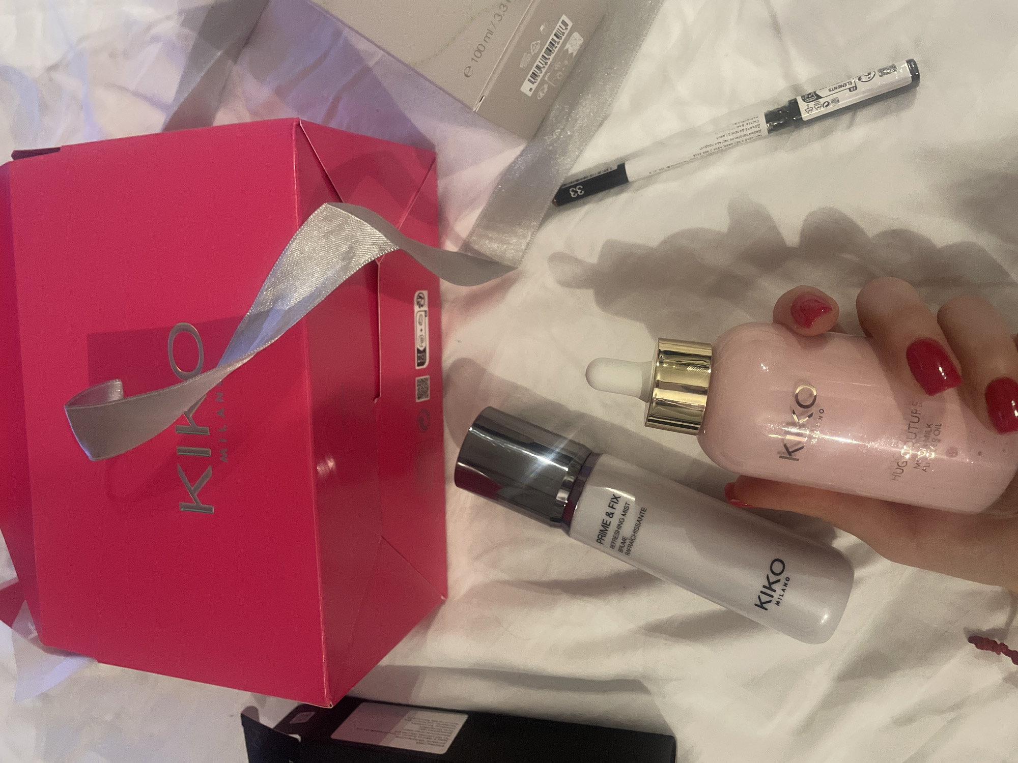 The scene is an indoor, domestic setting, specifically an unspecified location within the city of Sofia, Bulgaria. Multiple cosmetic products and their packaging are displayed on a white, rumpled fabric surface. A human hand, with red-painted fingernails, holds a light pink Kiko Milano "Hug Couture" bottle, identified as "BODY MILK ALOE OIL" with a gold-colored pump. Adjacent to it, a light grey Kiko Milano "Prime & Fix" refreshing mist bottle is positioned. A hot pink Kiko Milano branded gift box or shopping bag with silver lettering is centrally located, featuring a silver ribbon extending across the frame. To the right of the ribbon, a slender white and black cosmetic pencil, marked with "33" and "FLORENTS," lies flat. Additional packaging elements include a partial view of a black box on the upper left and a light grey box displaying "e 100 ml / 3.3 fl. oz." and a barcode on the upper right.