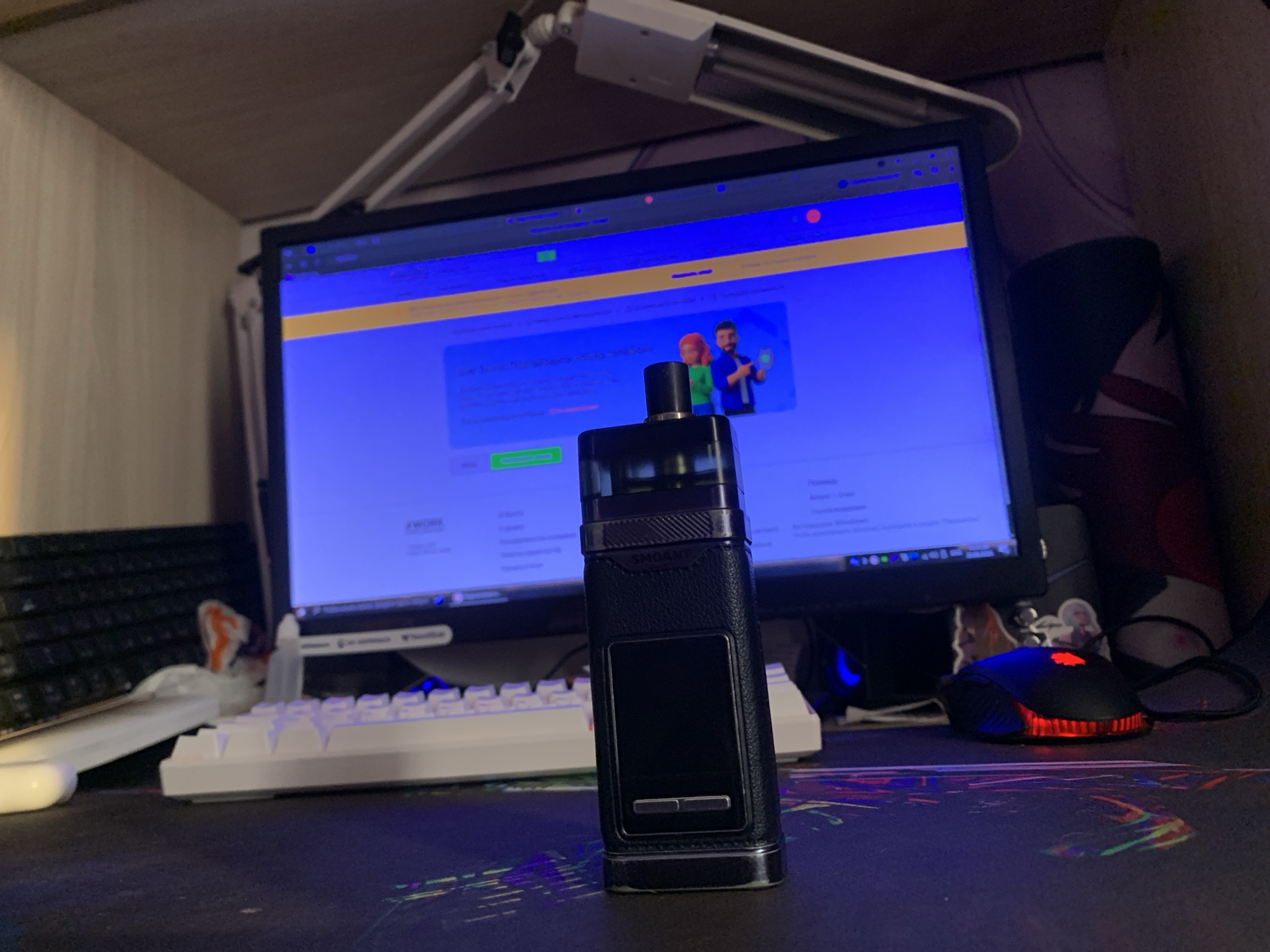 A desktop computer monitor displays a blue screen with yellow and white elements. In the foreground, a black electronic cigarette device stands upright. To the left of the device is a white keyboard, and further left is a black keyboard. A black computer mouse with red lighting is situated to the right of the electronic cigarette. The scene is set on a desk with a patterned surface. The overall environment appears to be an indoor workspace. There is no discernible indication of the city of Norilsk, Russia, or any specific location within it.