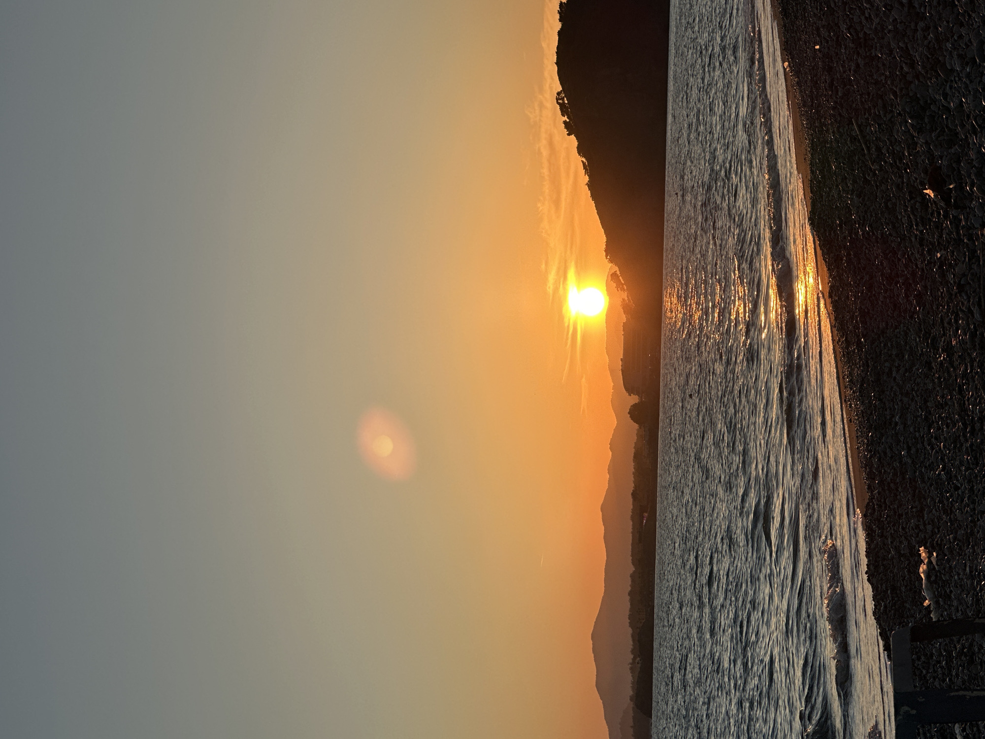 The image captures a sunset over the sea in Fethiye, Turkey. The sun is setting behind a mountain range, creating an orange glow in the sky. The sea is reflecting the sunlight, and waves can be seen crashing on the shore. The shore is a pebble beach, possibly Calis Beach, a popular spot to watch the sunset. No people are visible in the image.